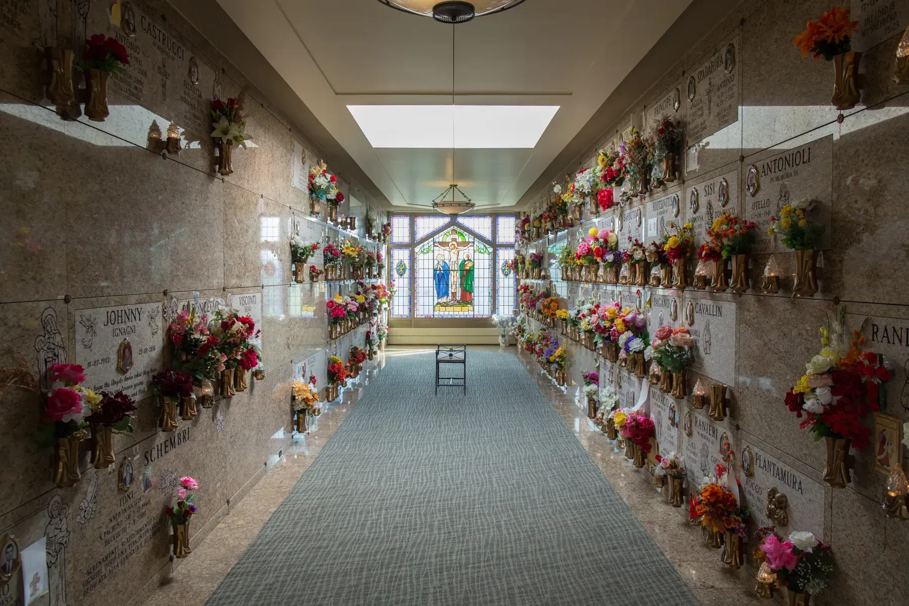 Flower-lined cemetery columbarium corridor with a bench and skylight at the end