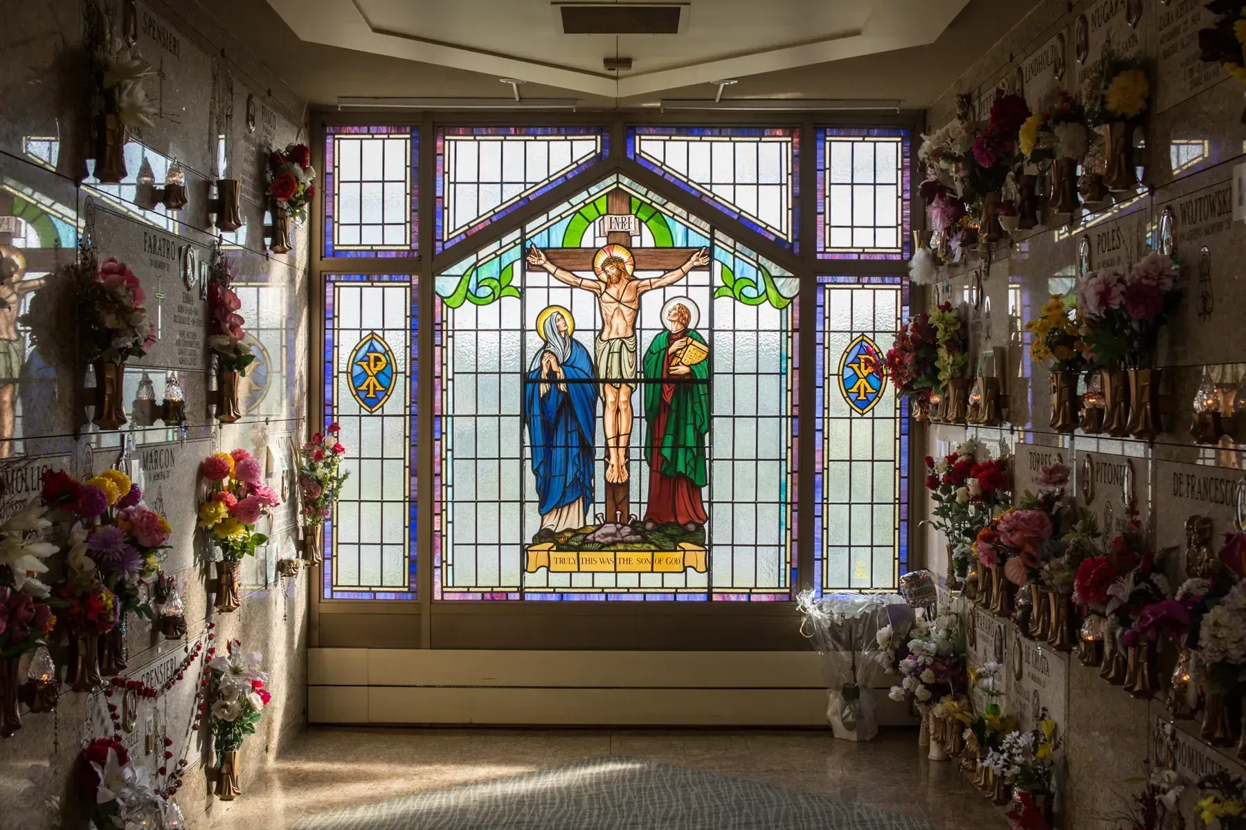 Stained-glass memorial display with flowers lining both walls in a narrow chapel-like corridor