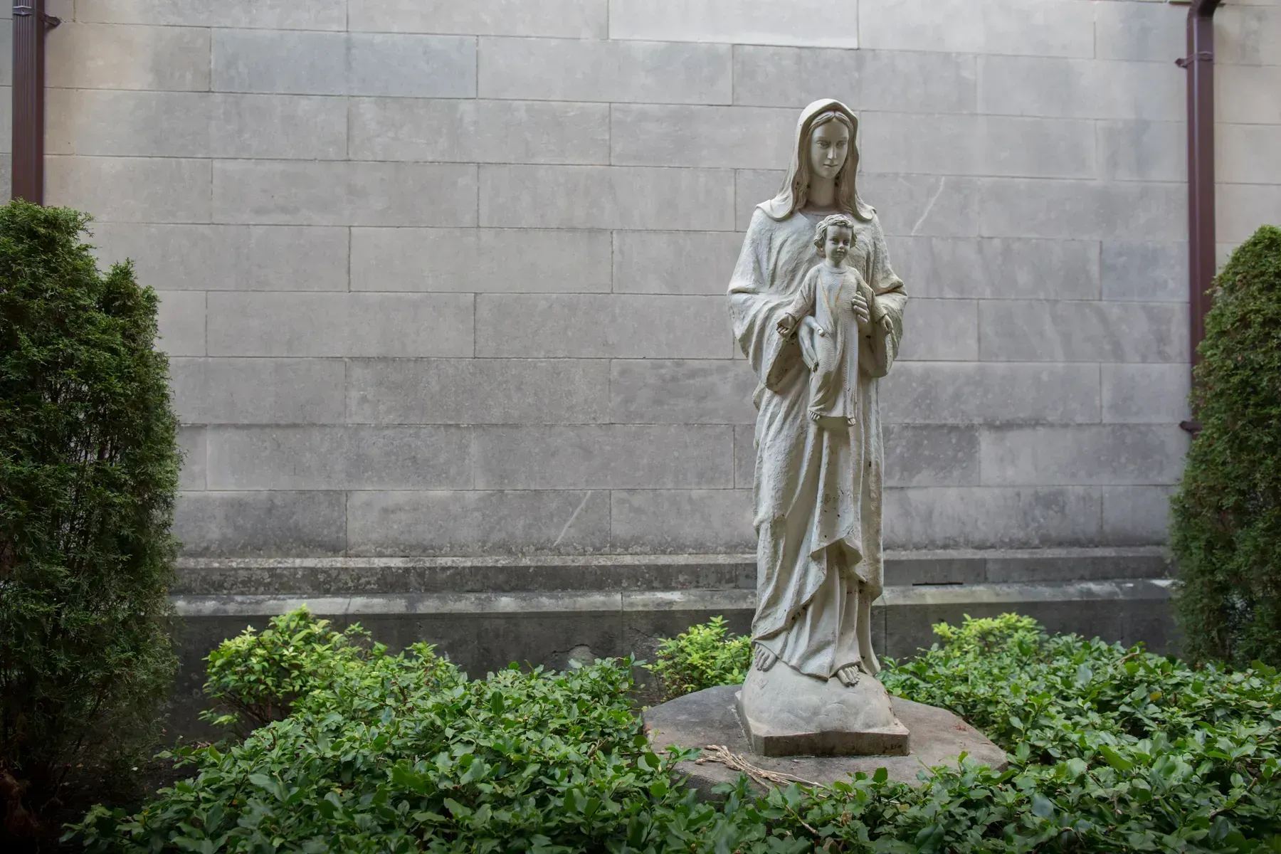 Stone statue of a robed woman holding a child, set in a garden against a gray wall.