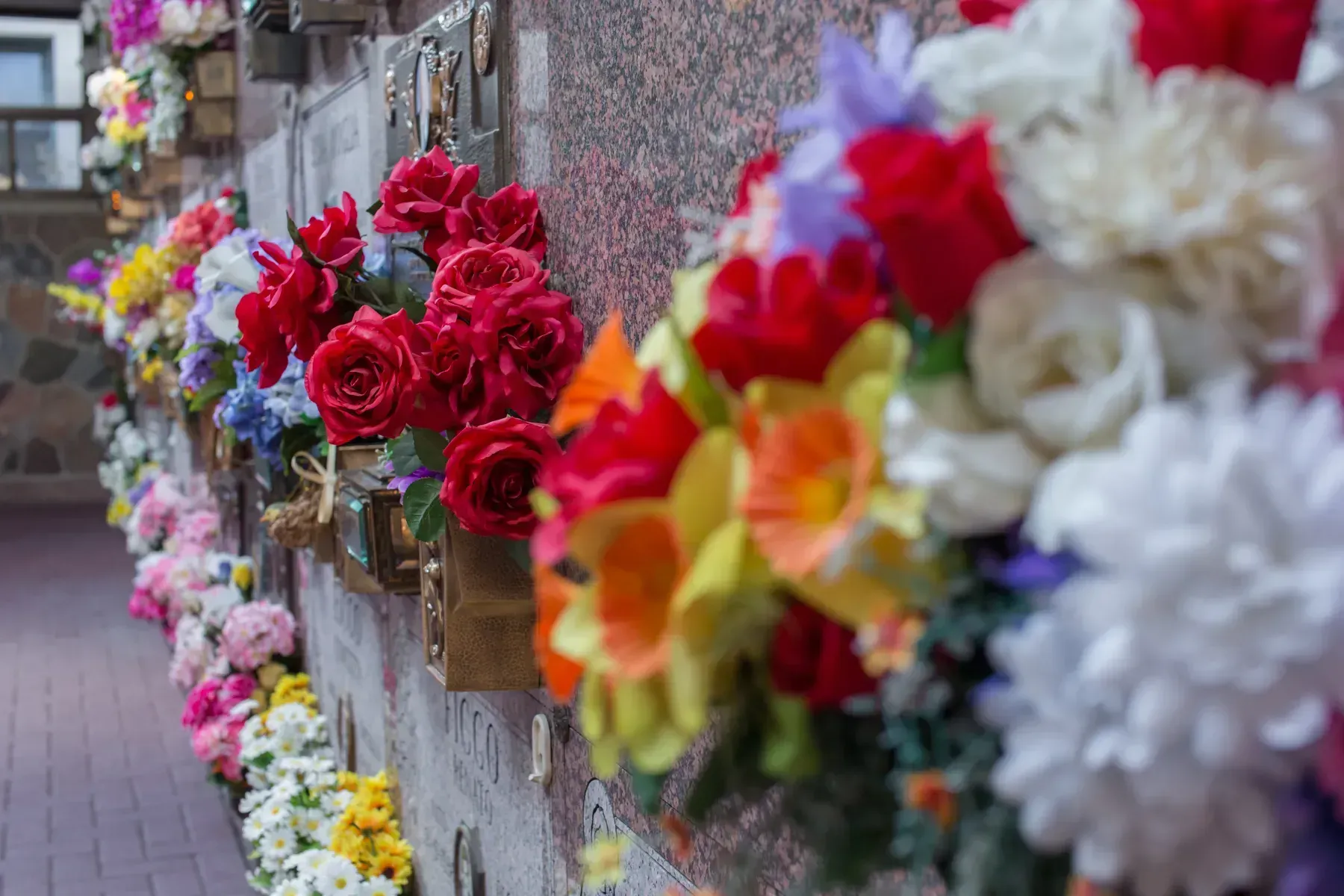 Flowers arranged along a stone memorial wall, with bright red, white, and yellow bouquets.