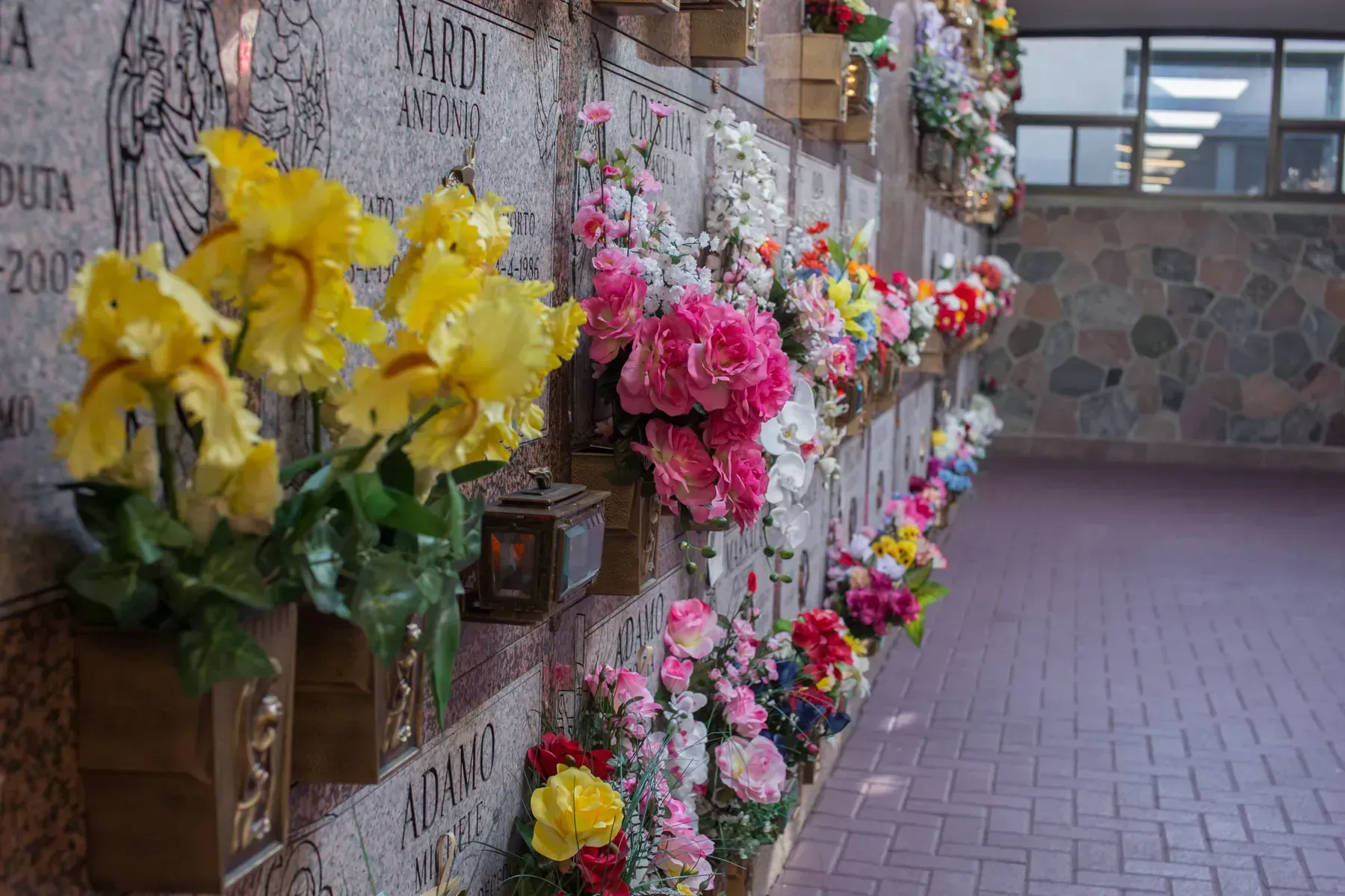 Flower-covered memorial wall with yellow and pink bouquets along a brick walkway