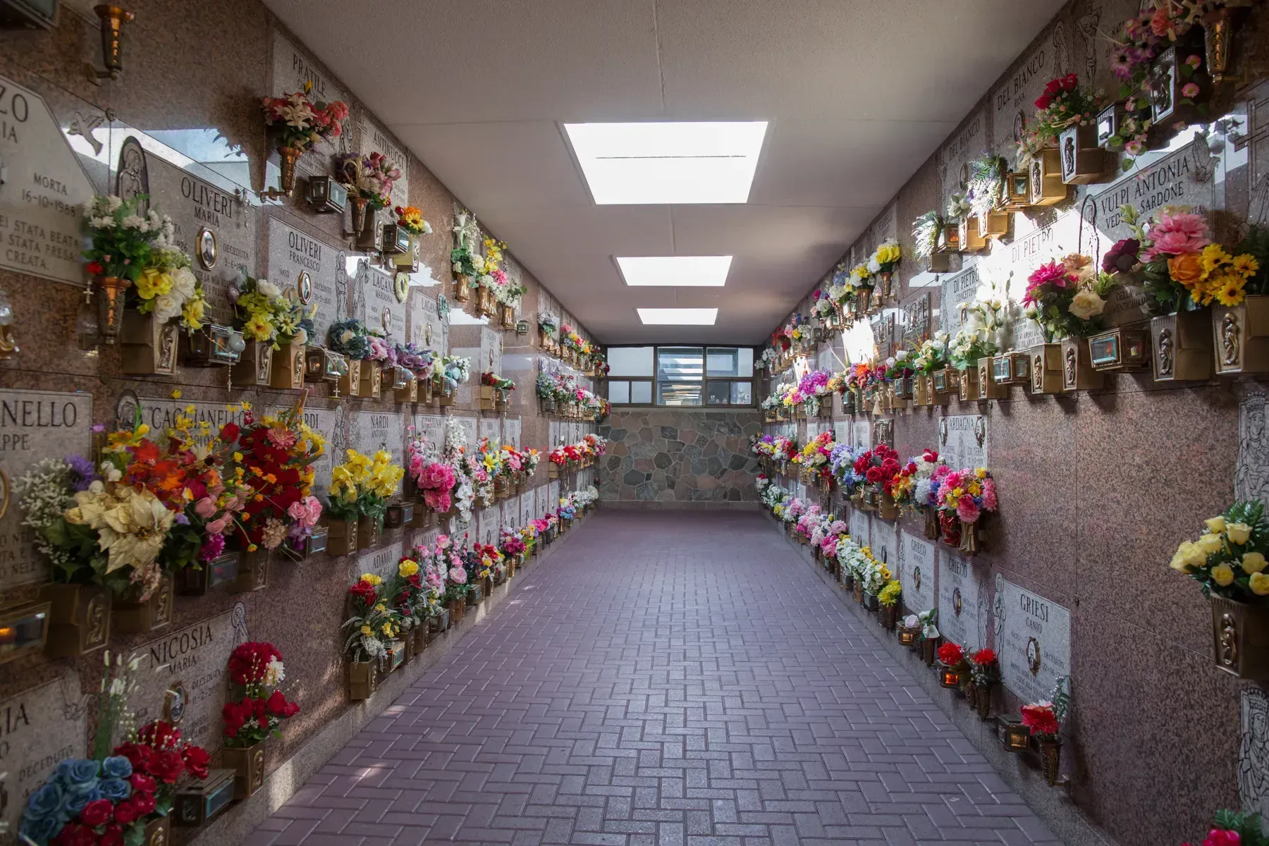 Indoor cemetery columbarium hallway lined with colorful flower arrangements and memorial plaques