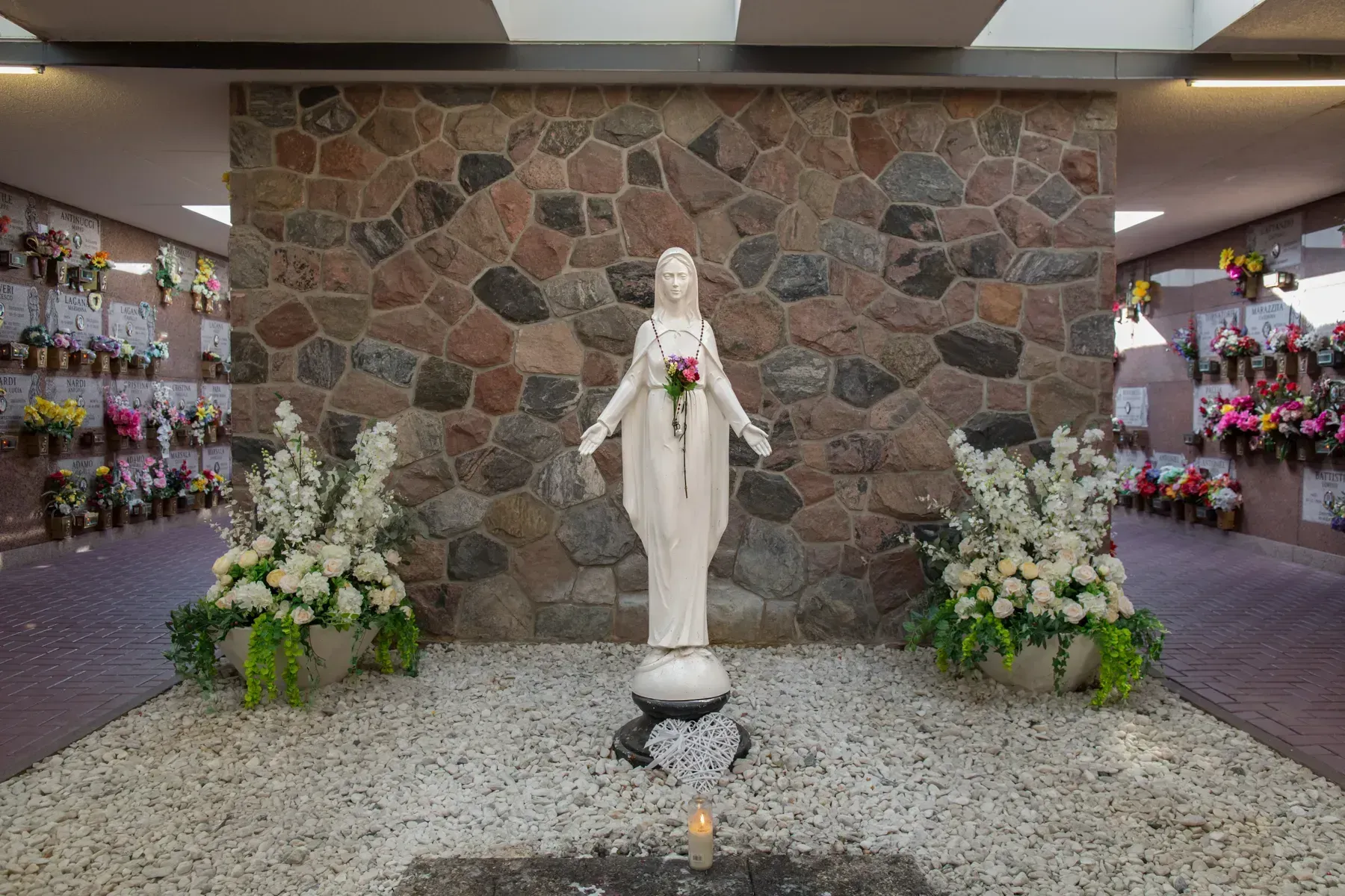 White statue in a flower-lined memorial alcove before a stone wall, with candles and floral arrangements.