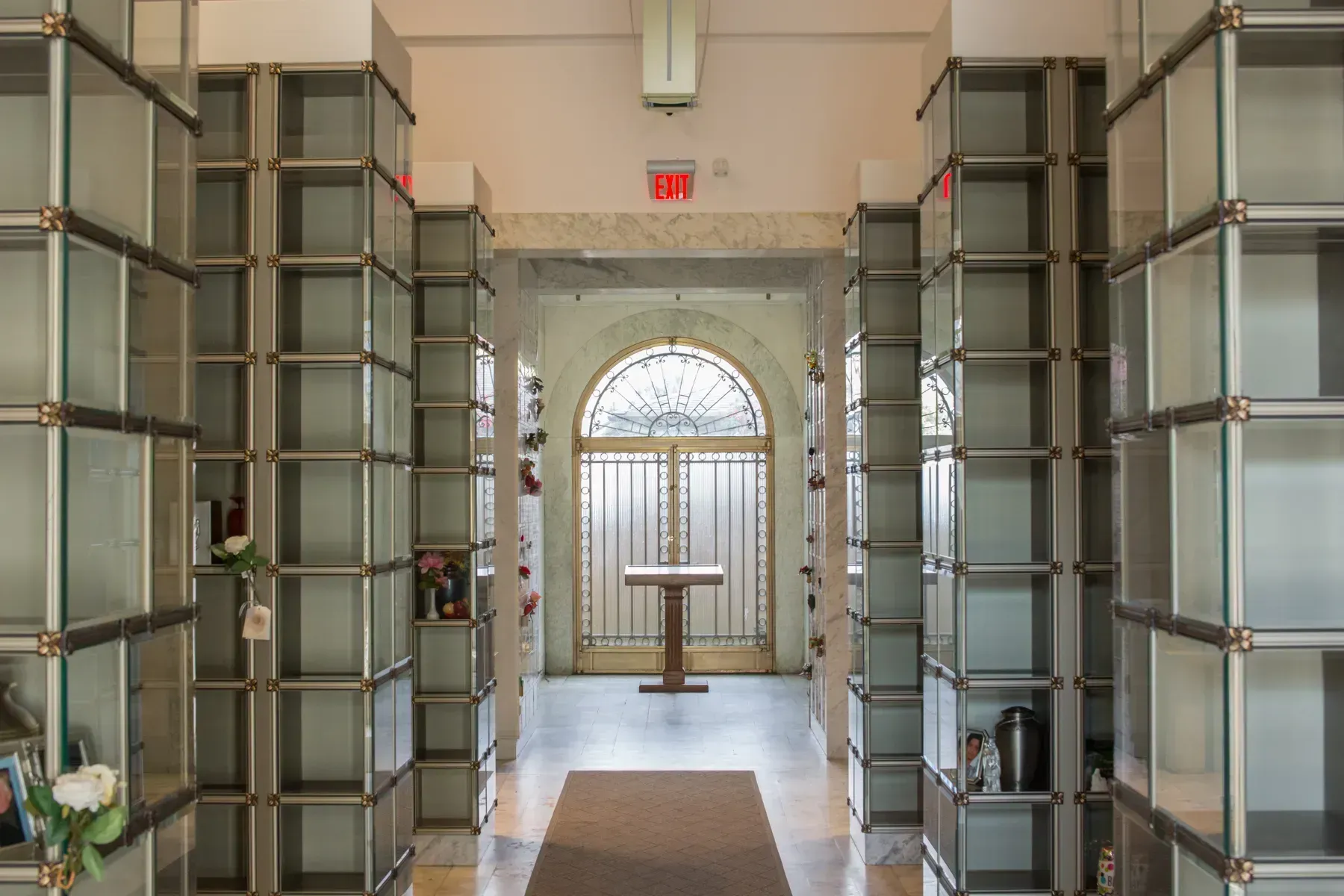 Narrow mausoleum corridor with glass-front crypt niches and an arched stained-glass window at the end