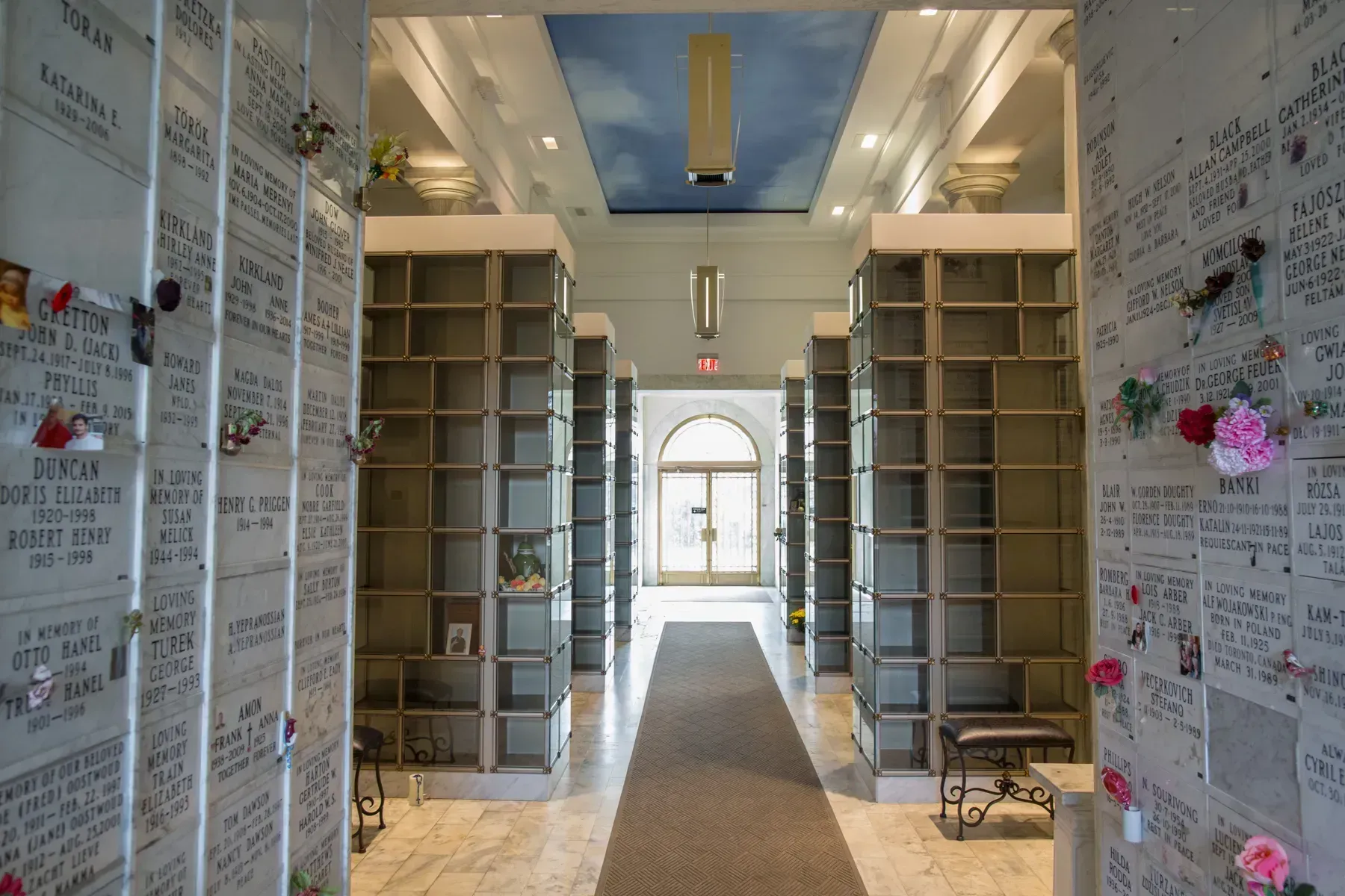 Narrow aisle between tall shelves, leading to a bright arched doorway in a white, note-covered room
