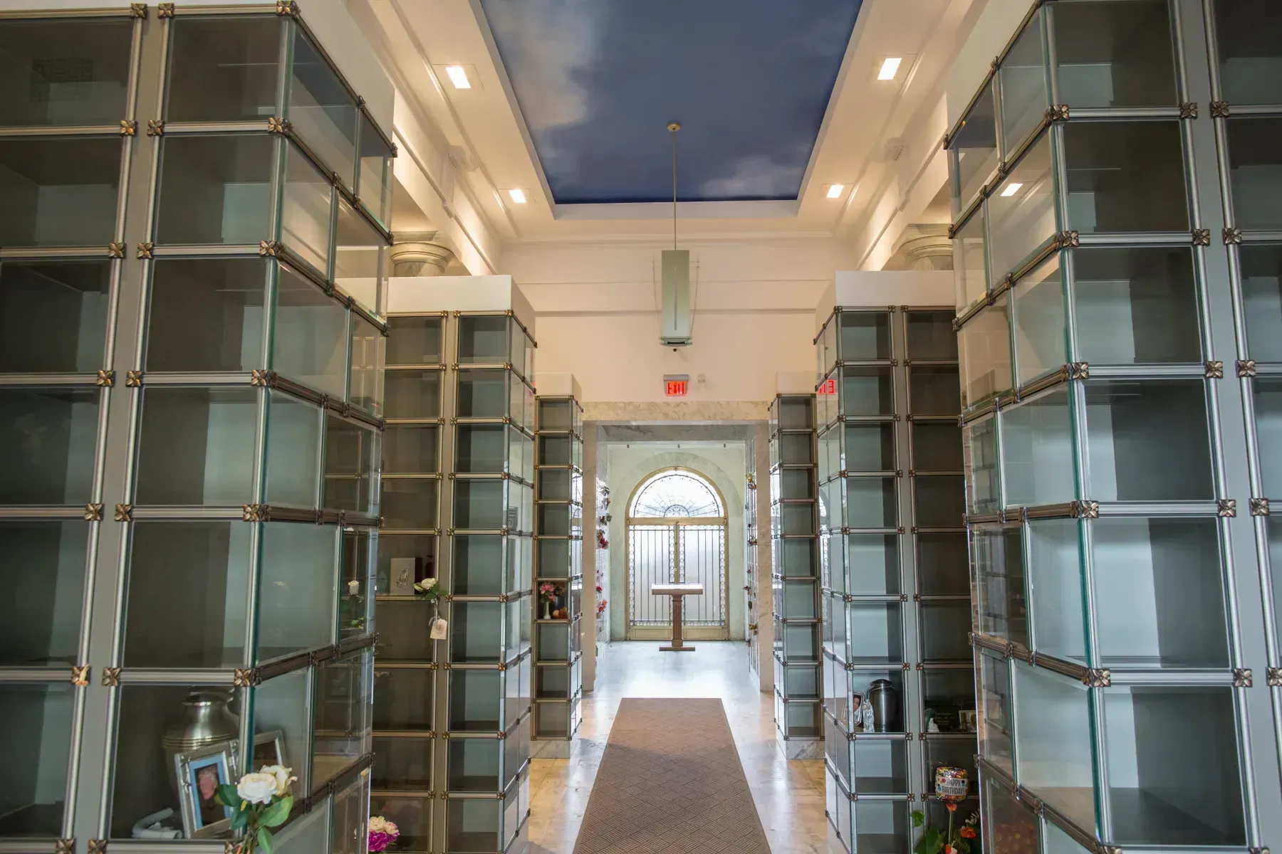 Bright hallway lined with glass display shelves, leading to a sunlit doorway and blue sky beyond