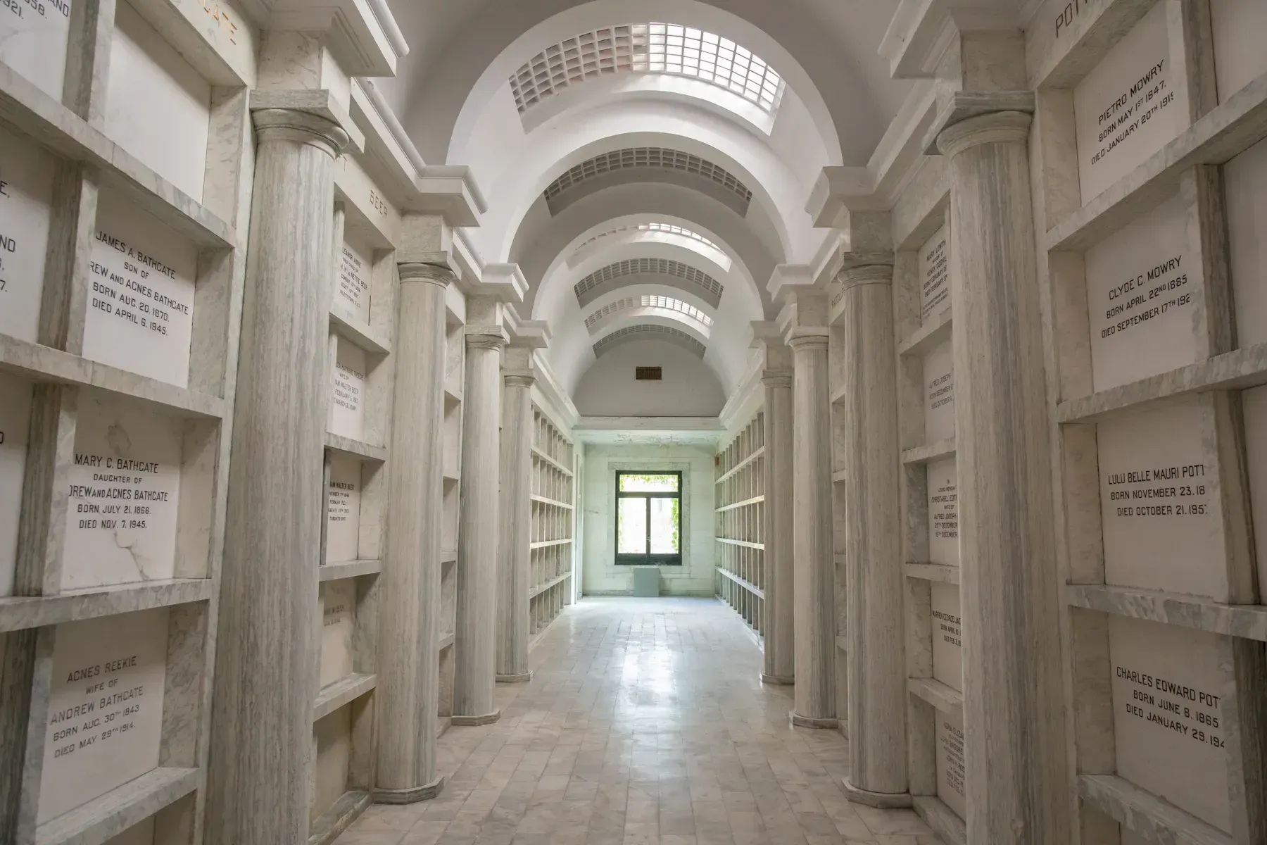 White mausoleum corridor with arched ceilings and niches, lit by a bright window at the end