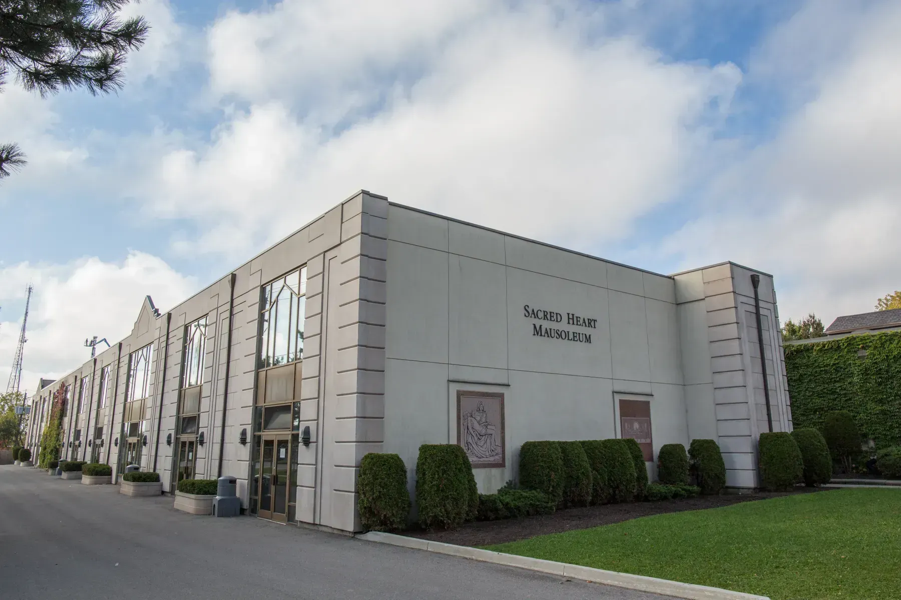 Modern white building with tall windows, trimmed hedges, and a cloudy sky overhead