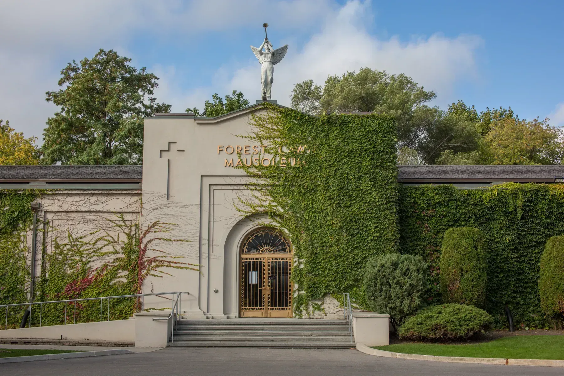 Stone chapel entrance with ivy-covered wall, arched doorway, and cross on the roof under a blue sky