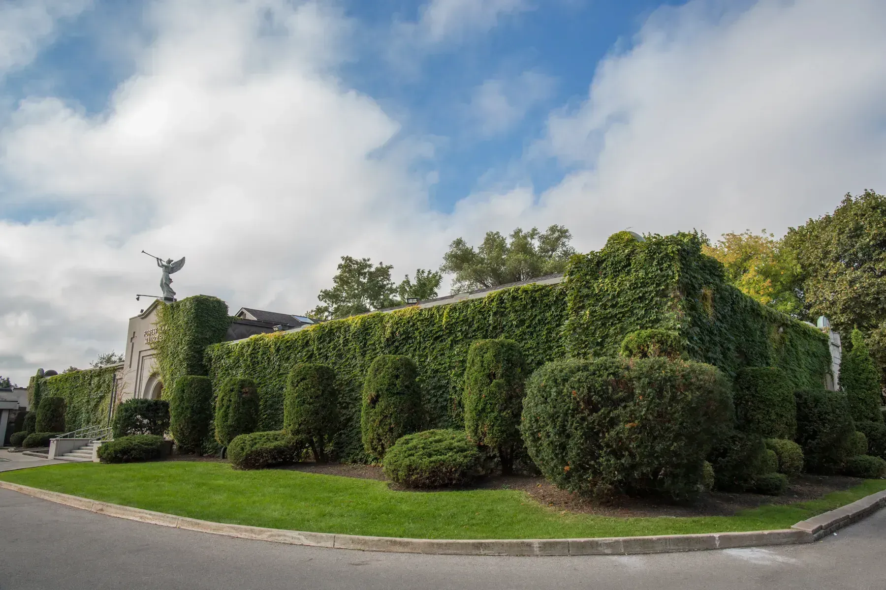 Brick building covered in green ivy beside a road, with manicured bushes and cloudy blue sky