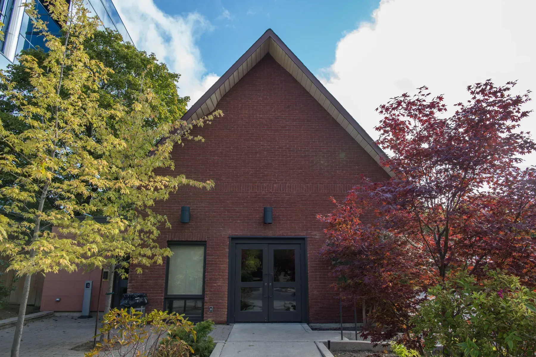 Red brick building entrance with dark double doors, framed by green and red trees.