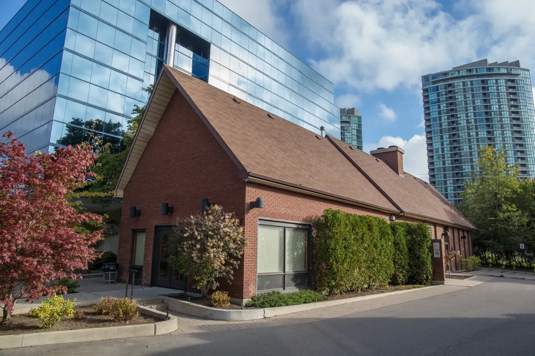 Brick building with steep roof beside a street, trees, and modern high-rise towers in the background