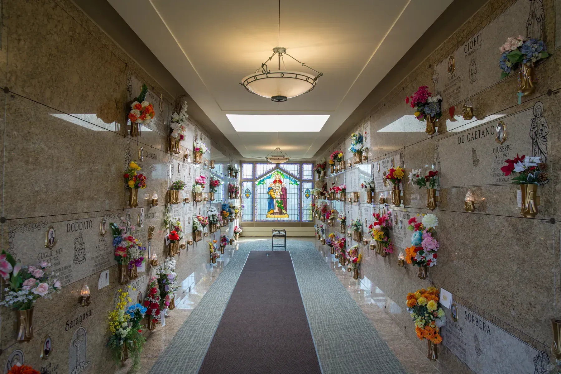 Flower-lined mausoleum hallway with a central carpet and stained-glass window at the end