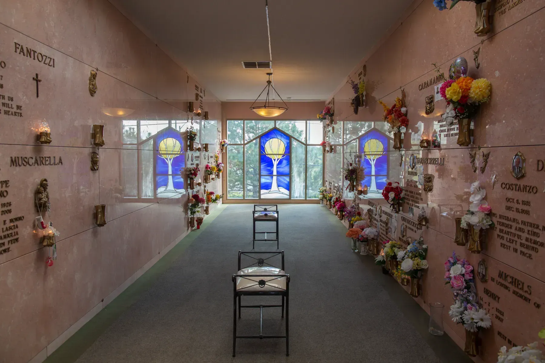 Narrow mausoleum hall with flowered memorial niches and stained-glass windows at the end.