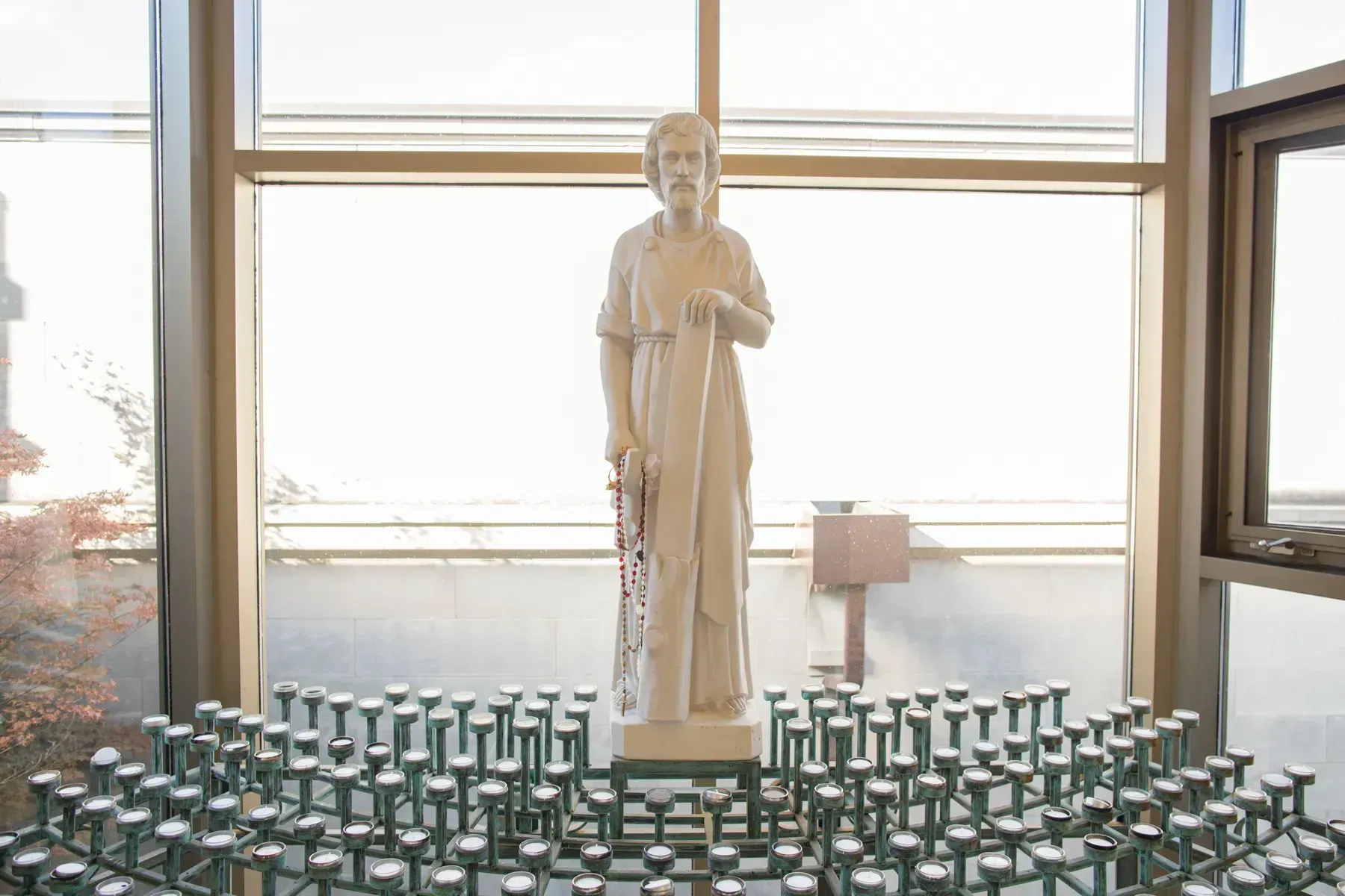 White statue centered before large windows, surrounded by rows of green glass candles.