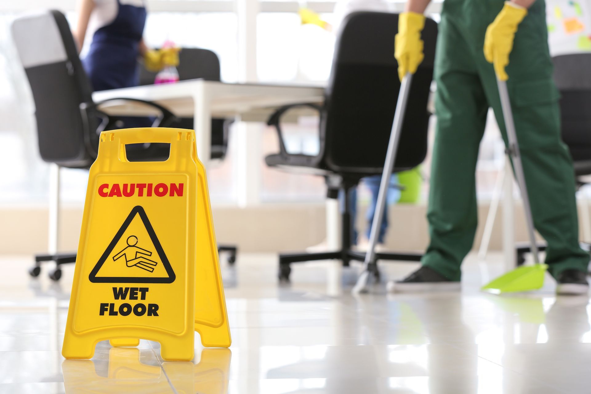 Yellow caution sign warning of wet floor in an office cleaning area with a mop and cleaner in the background