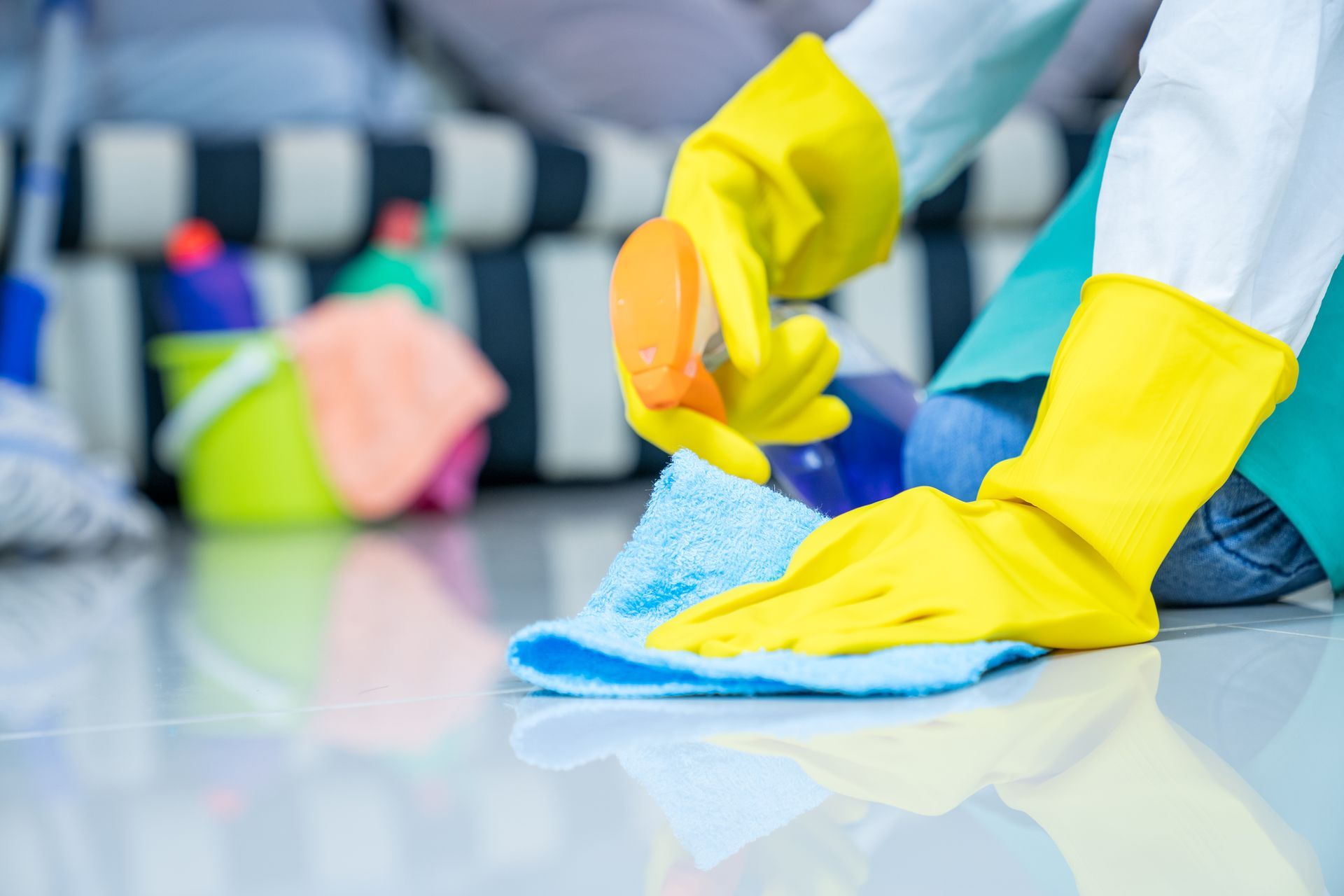 Gloved hands wiping a floor with a blue cloth and yellow spray bottle in a bright room