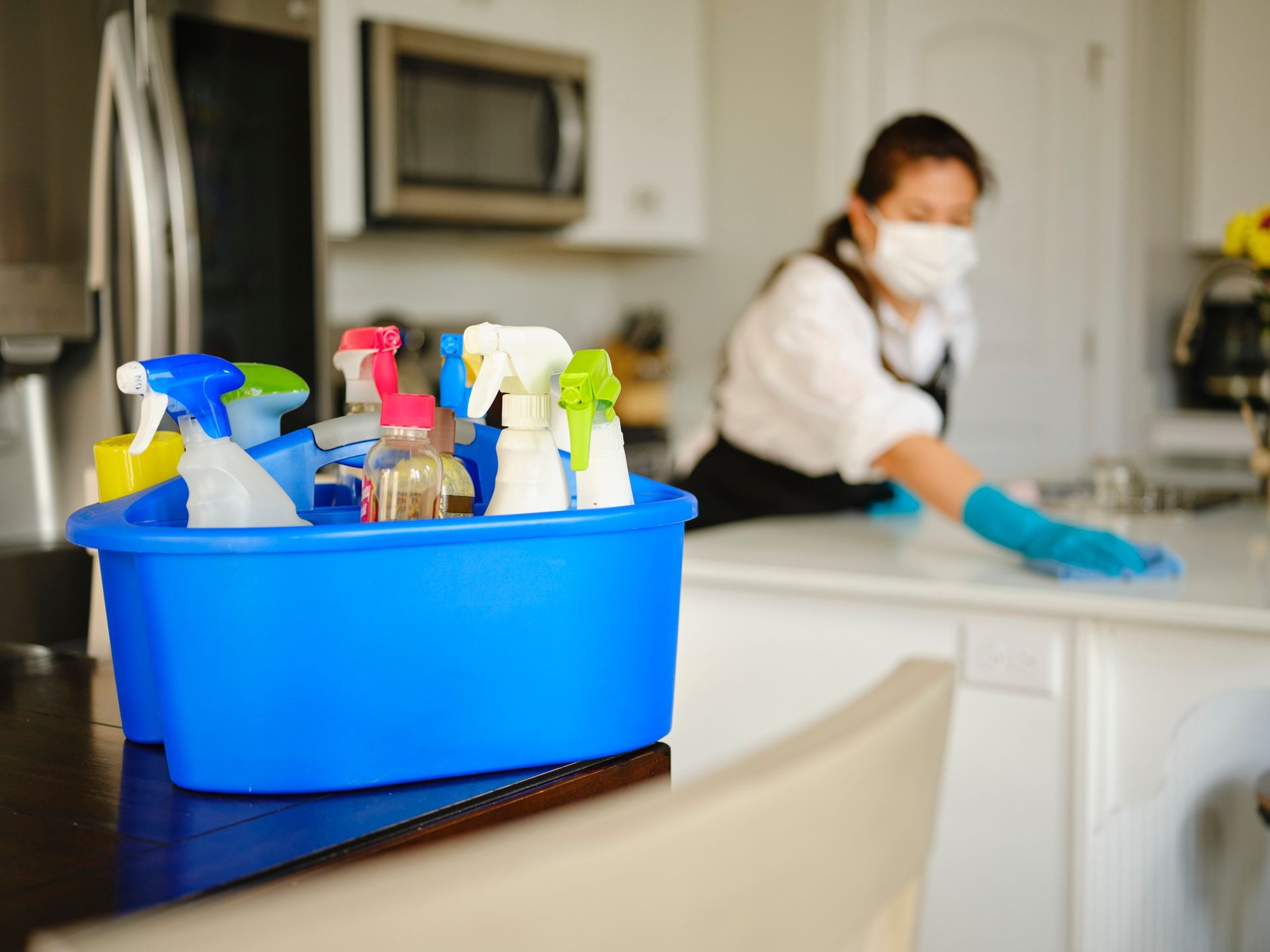 Blue caddy of cleaning sprays on a counter, with a masked cleaner wiping a kitchen in the background