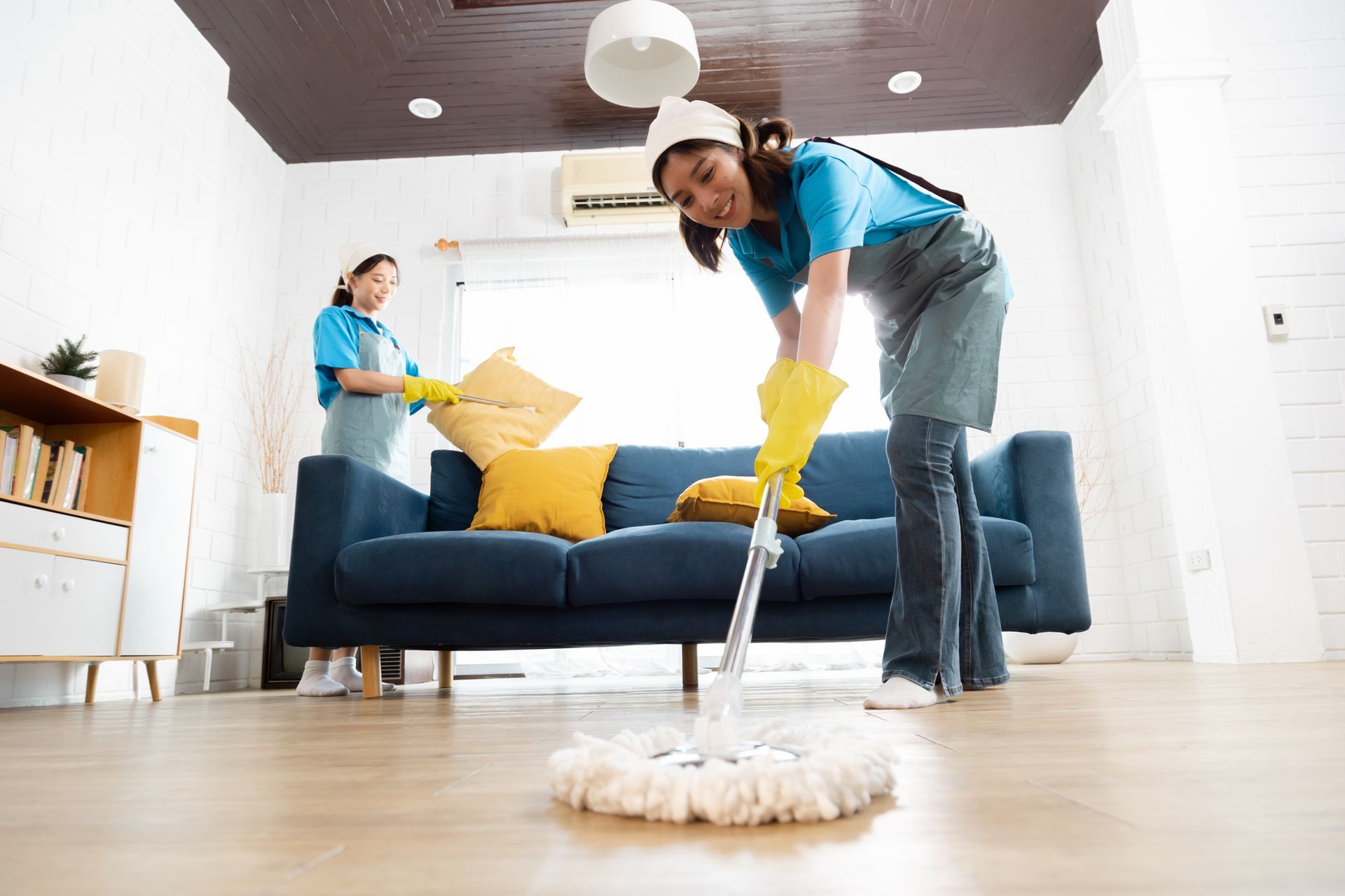 Two cleaners dusting and mopping a bright living room with a blue sofa and yellow pillows