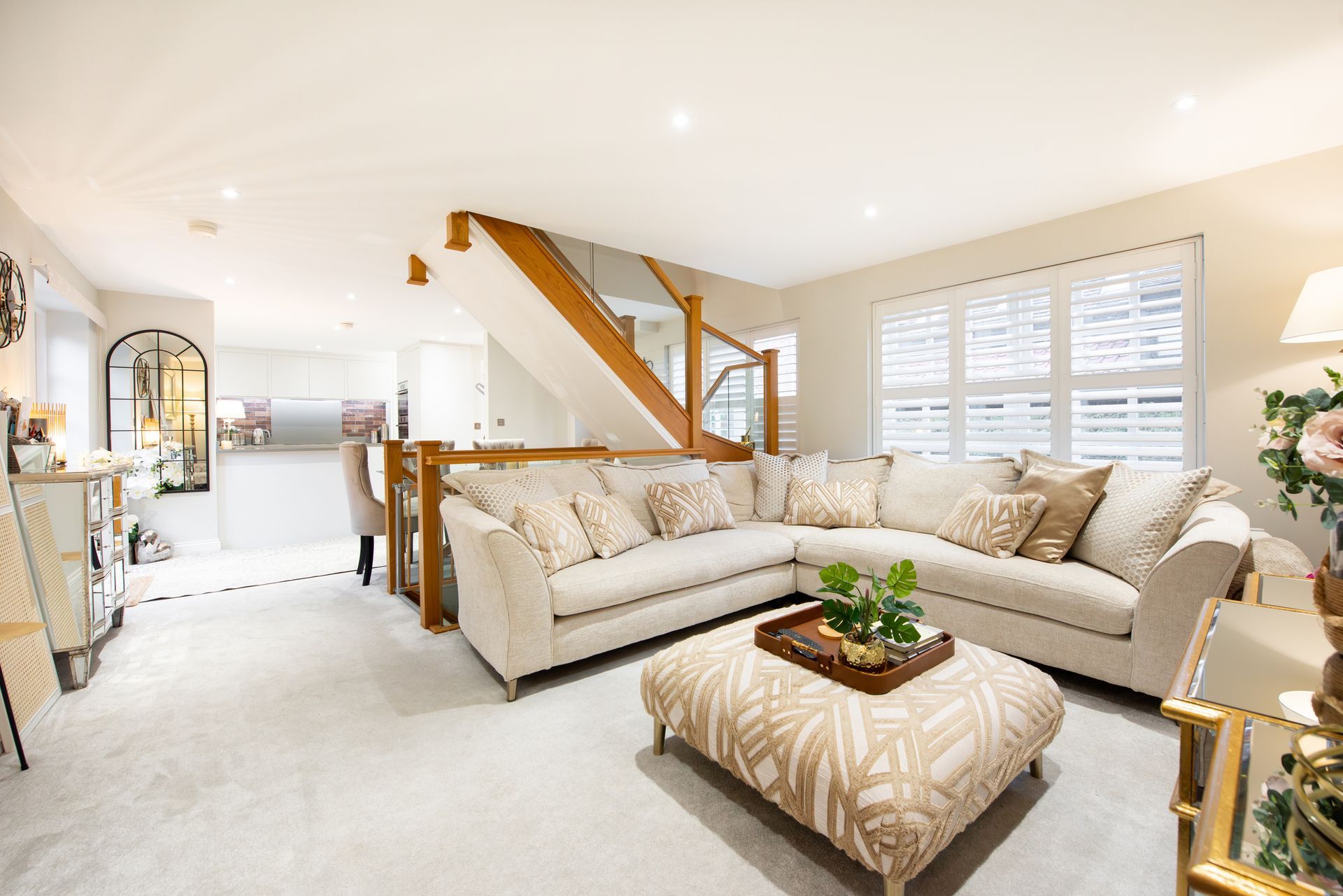 Bright open living room with beige sectional, ottoman, staircase, and sunlit windows