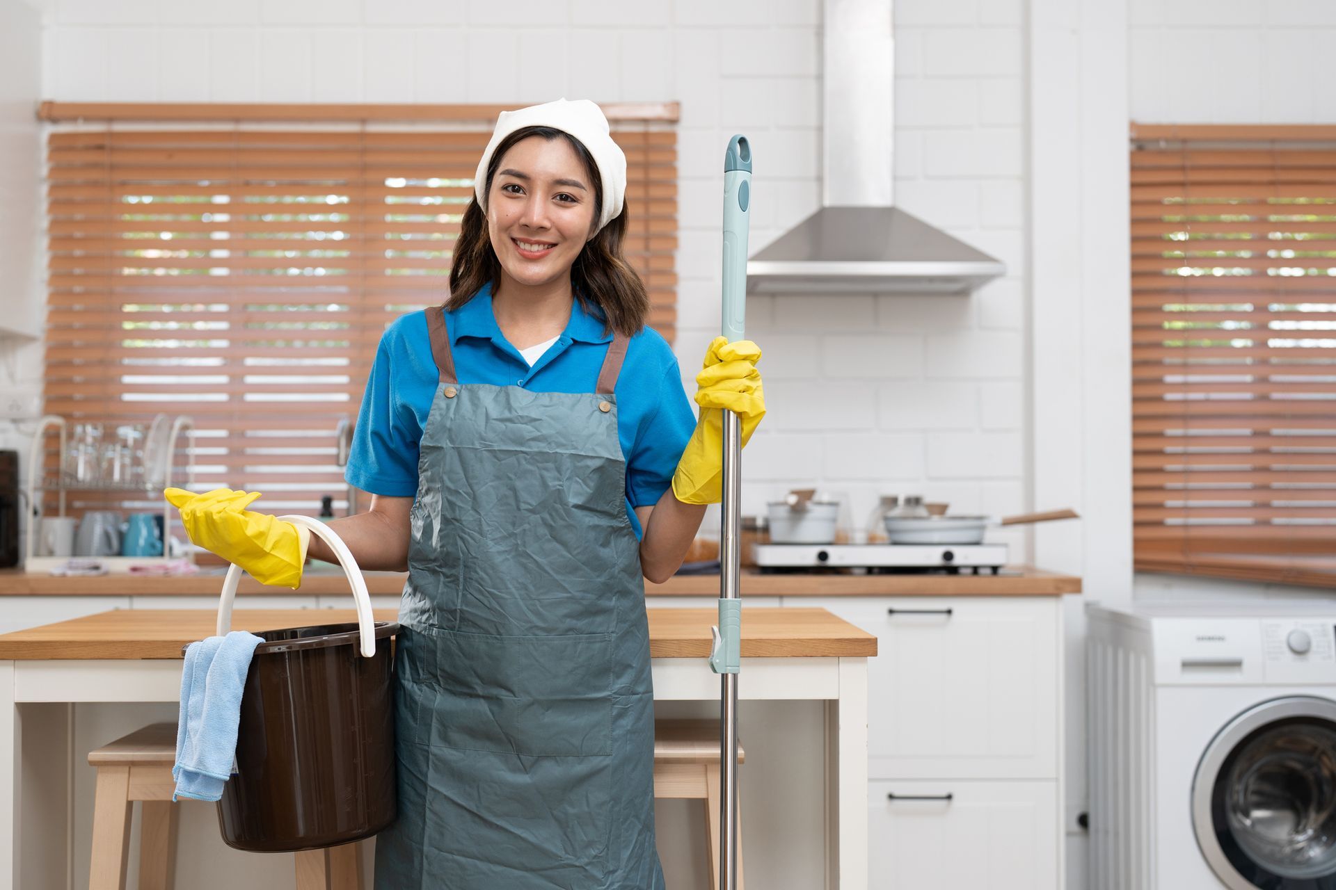 Cleaner in apron and gloves smiling in a kitchen, holding cleaning supplies beside a sink and washing machine.