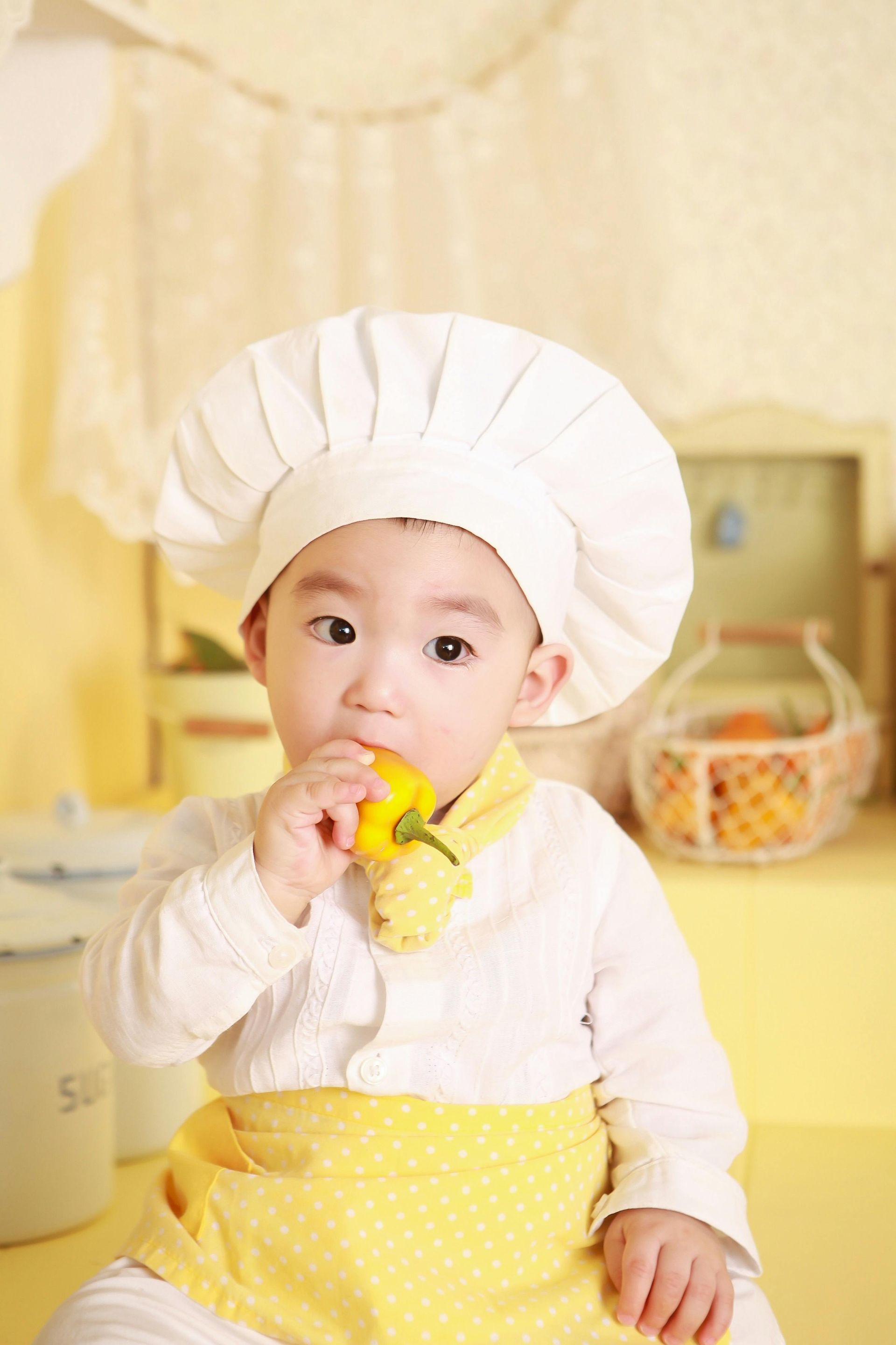 A baby wearing a chef 's hat and apron is eating a yellow flower.