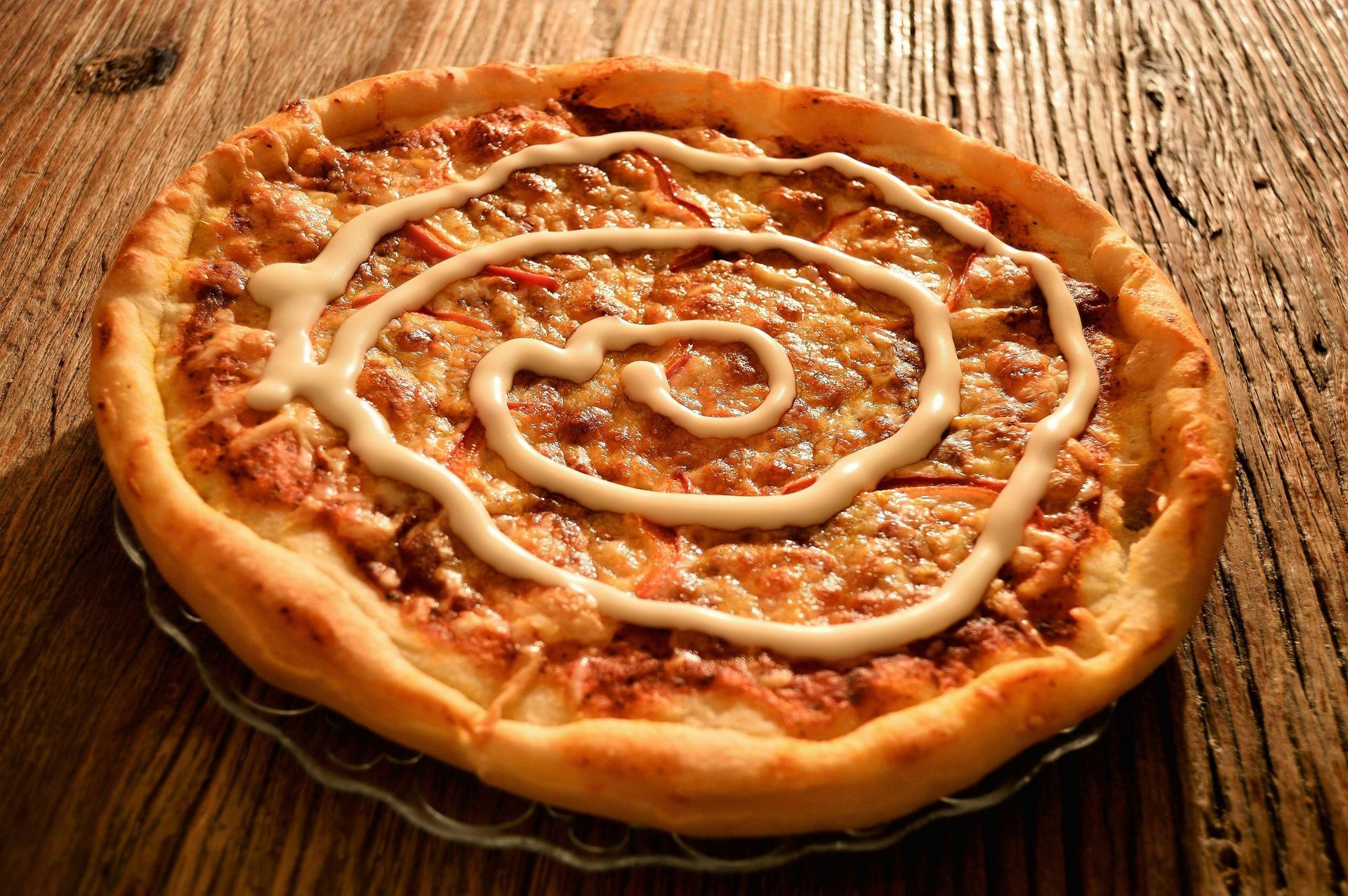 A pizza with a swirl of mayonnaise on top is on a glass plate on a wooden table.