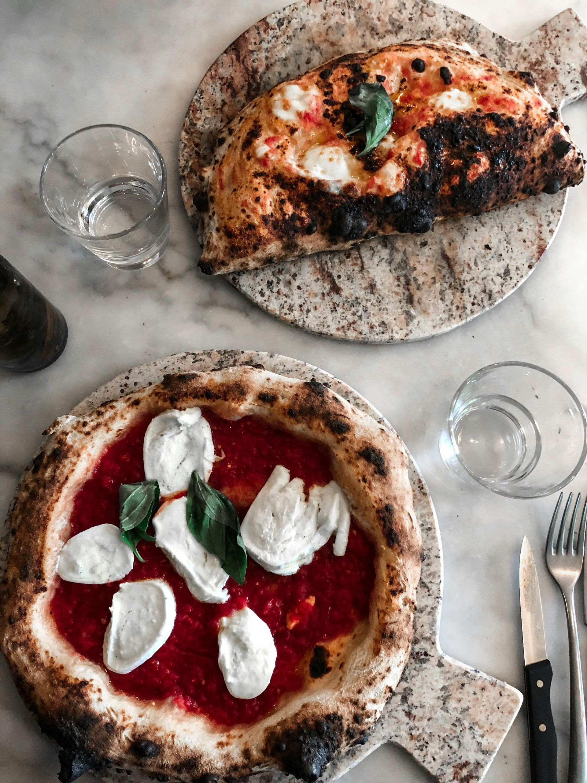 Two pizzas are sitting on a table with glasses and utensils