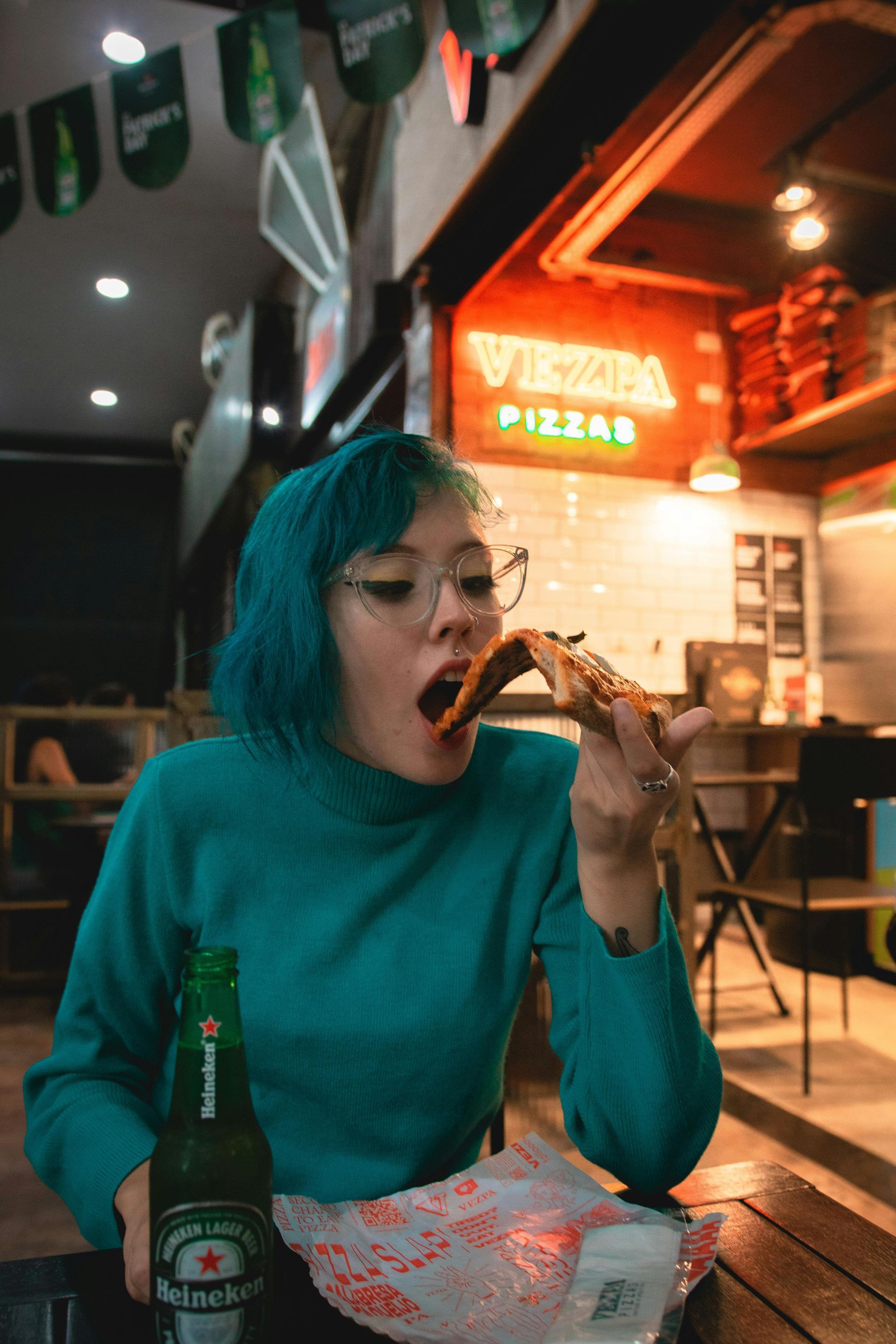 A woman with blue hair is sitting at a table eating pizza and drinking beer.