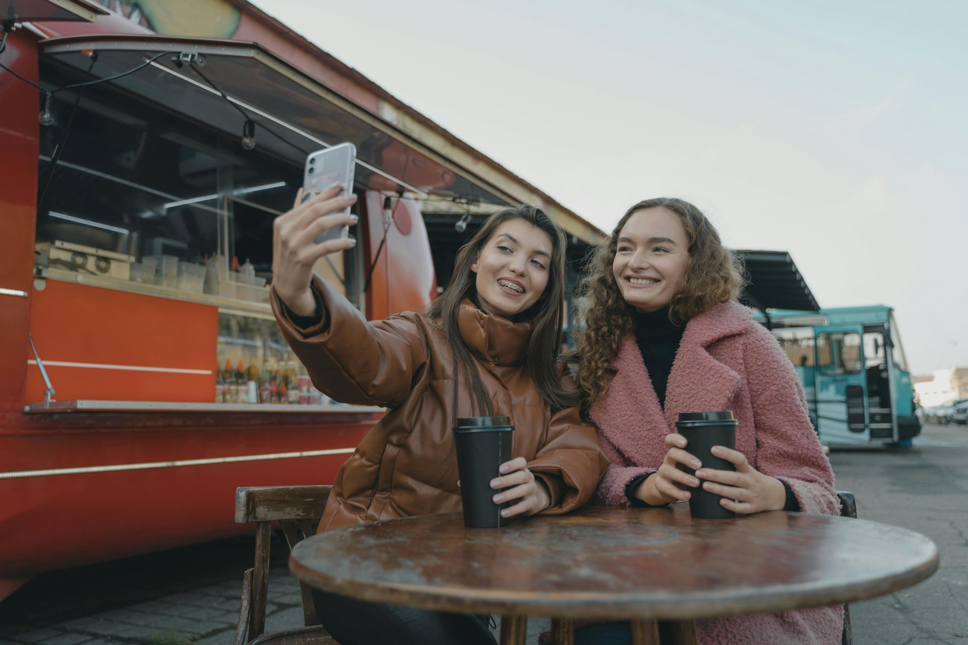 Two women are sitting at a table taking a selfie in front of a food truck.