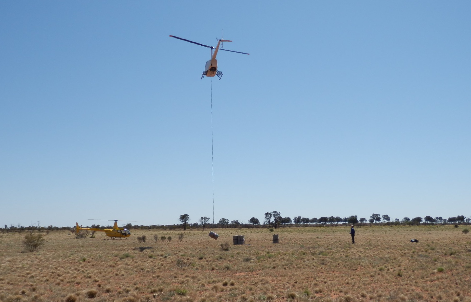 Helicopter flying over field, towing sensor. Another helicopter and person visible on the ground. Blue sky.