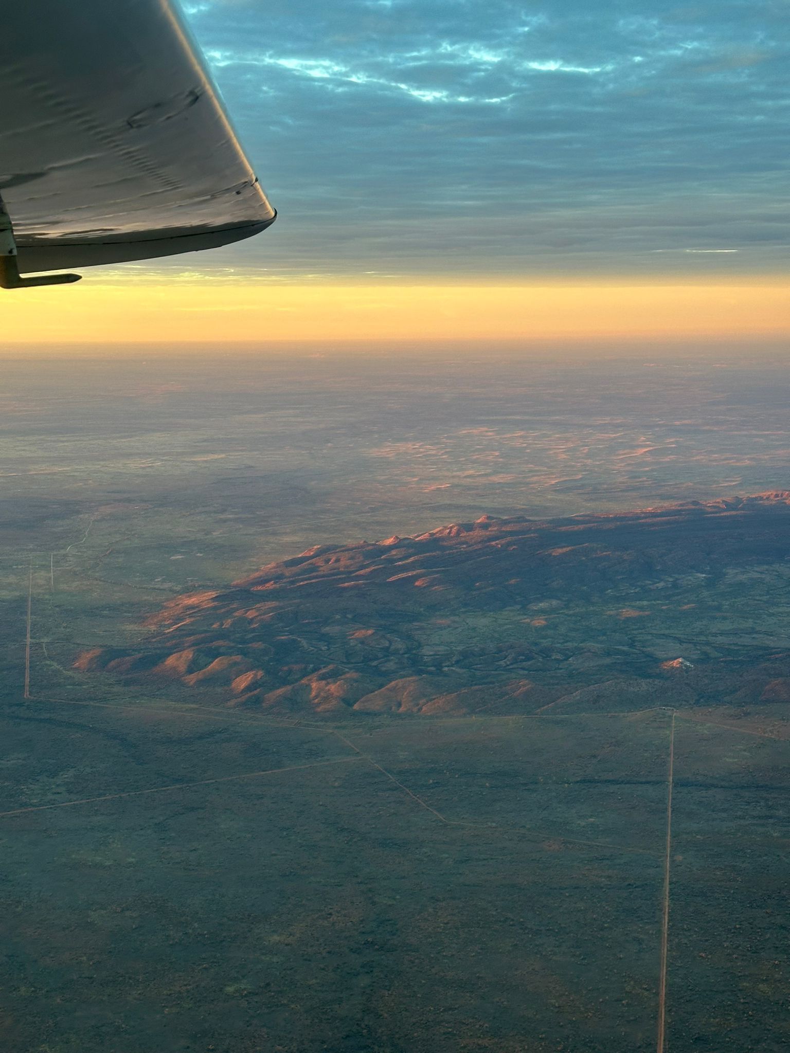 View from an airplane window of a mountainous landscape at sunset, with warm hues.