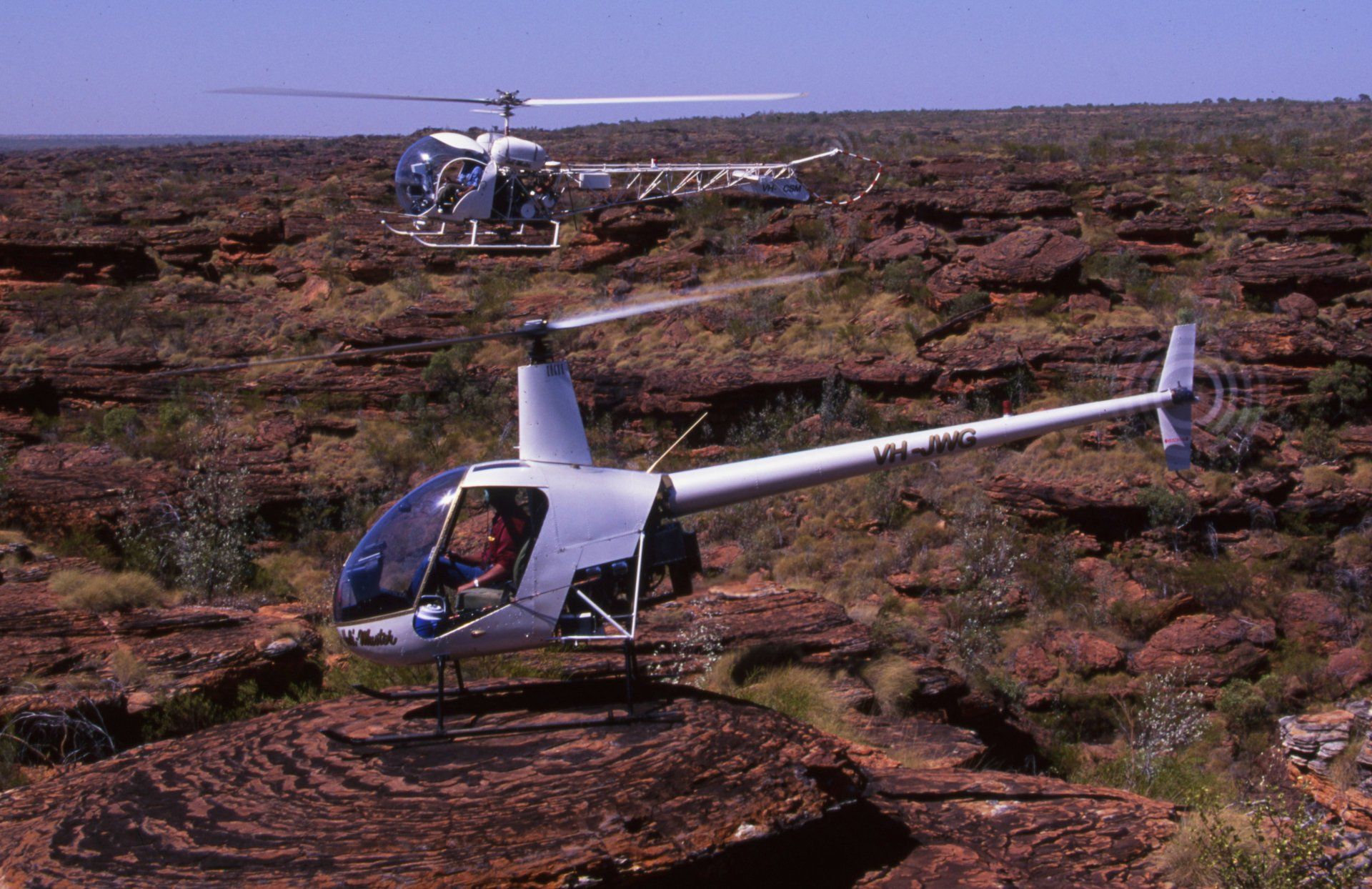 Two helicopters land on a reddish-brown, rocky terrain with sparse vegetation.