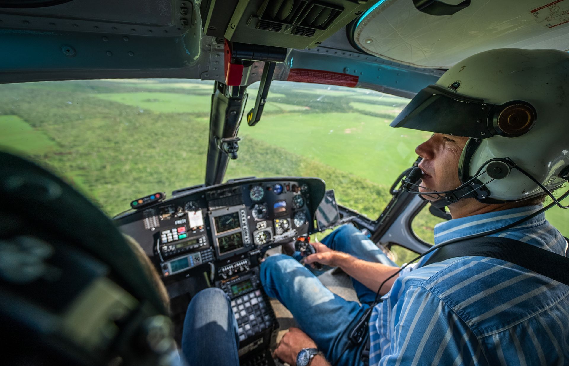 Pilot in helicopter cockpit, looking out window over green fields, wearing a helmet.