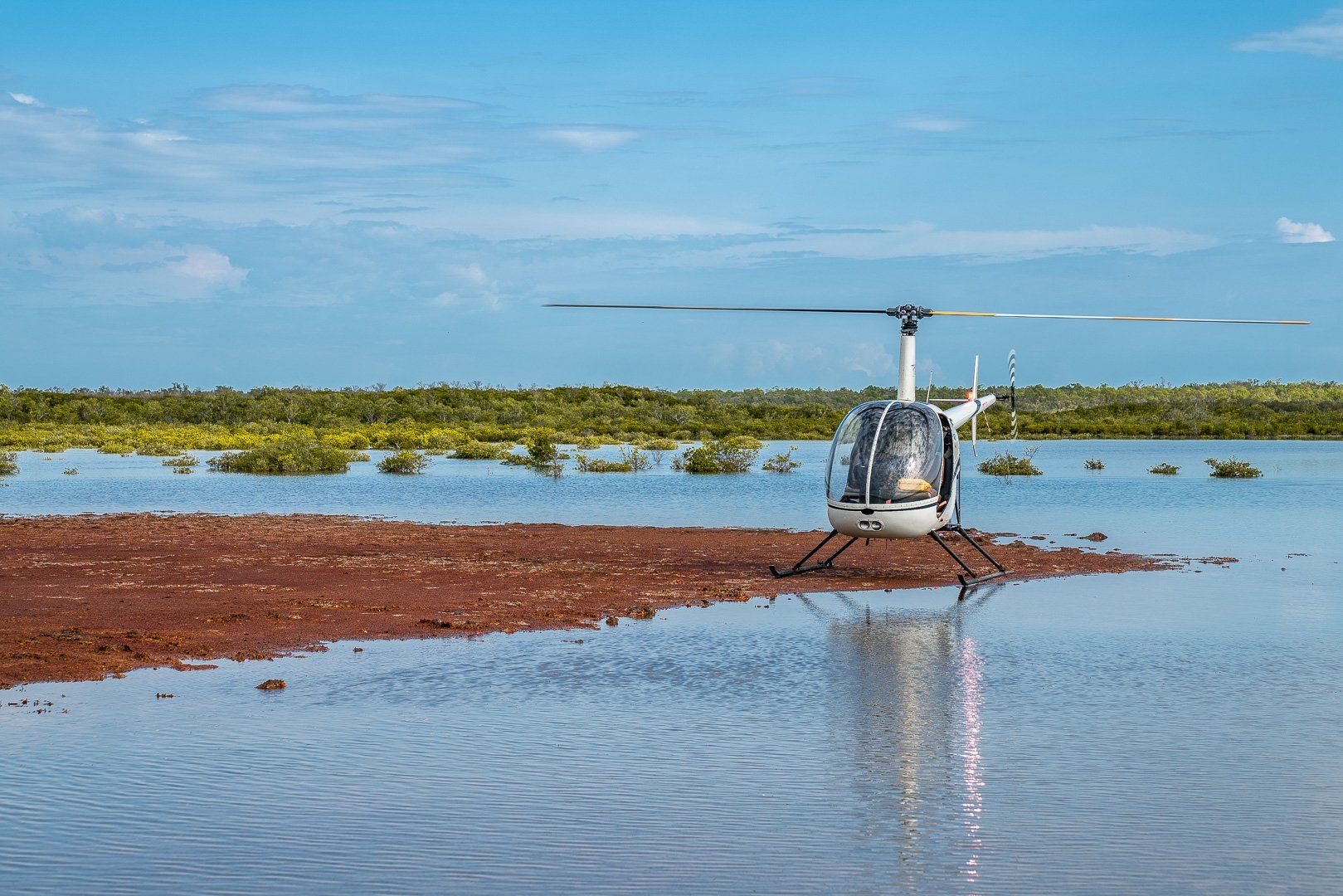 Helicopter parked on a small, reddish island in shallow water with a backdrop of green vegetation and a blue sky.