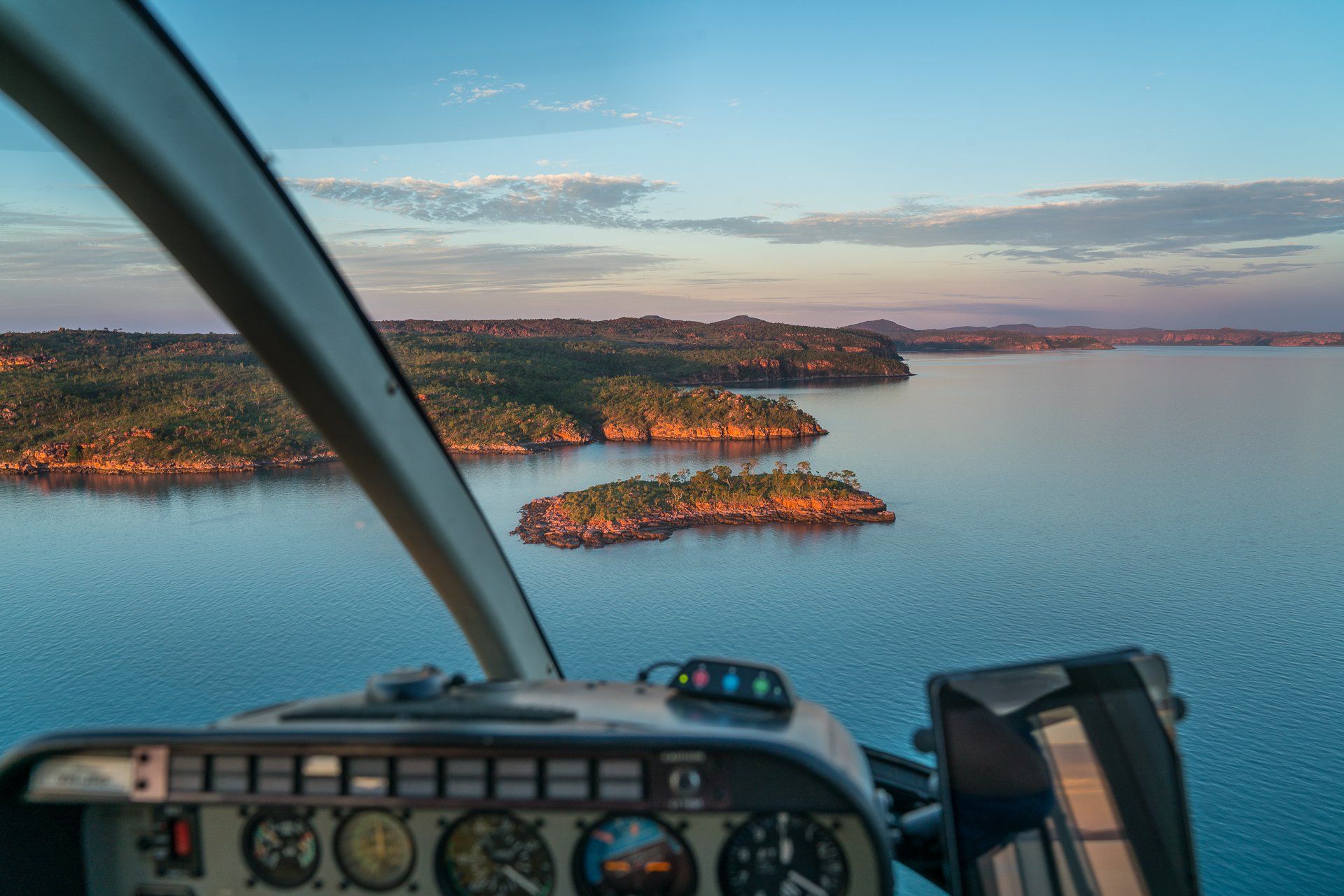 View from a helicopter cockpit: islands dot a blue lake at sunset.