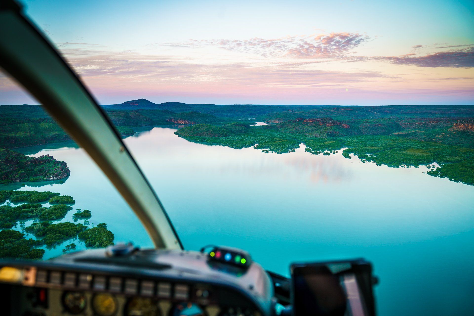 View from helicopter cockpit over a calm river and green landscape at sunset.