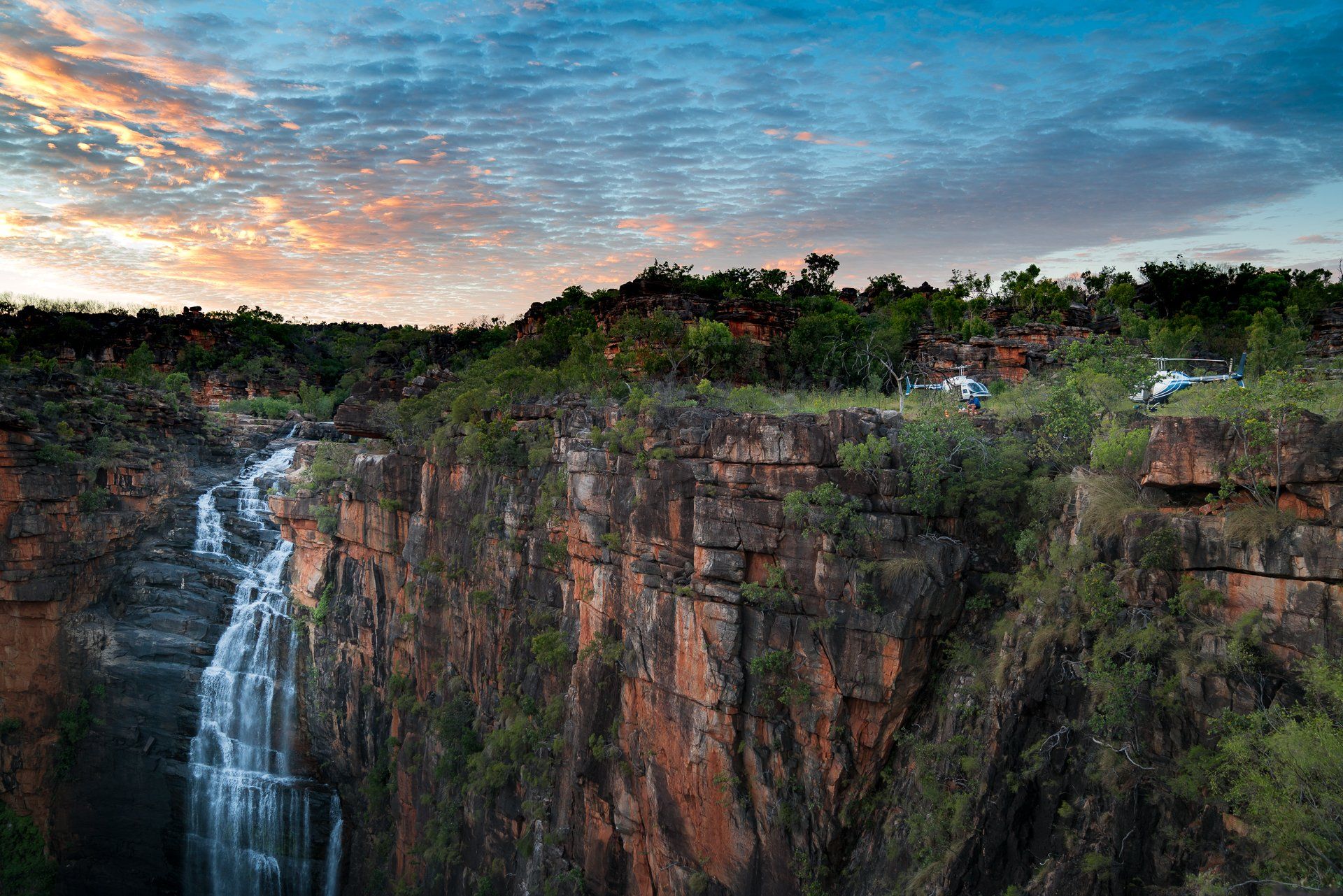 Waterfall cascading down a red rock cliff, lush green vegetation, with a sunset sky.