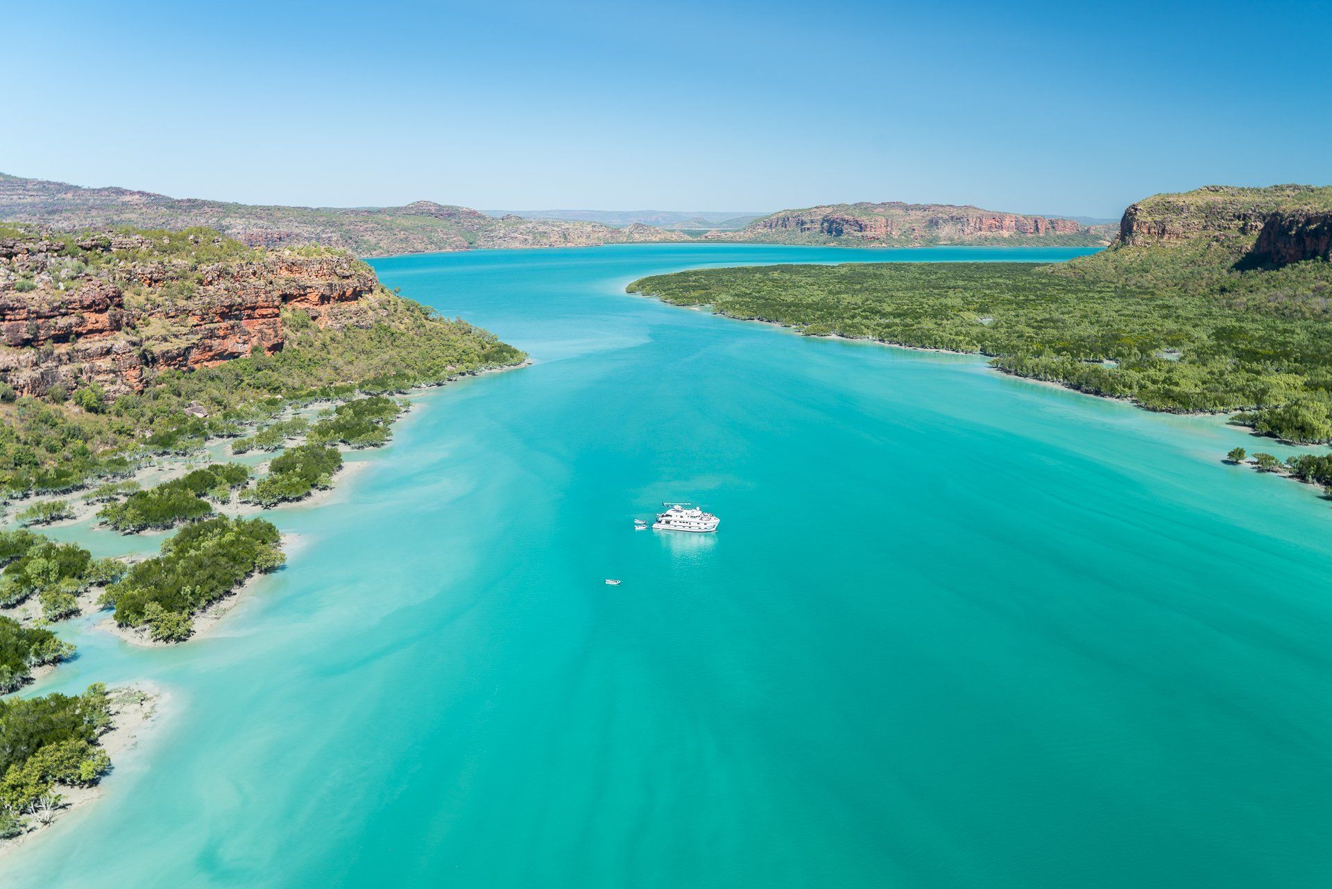 Turquoise water cuts through a landscape of red rock cliffs and green foliage with a white boat at center.