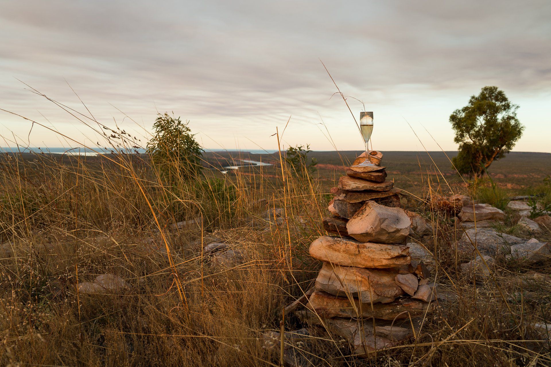 Stone cairn with glass balanced on top, overlooking a dry, grassy landscape at sunset.
