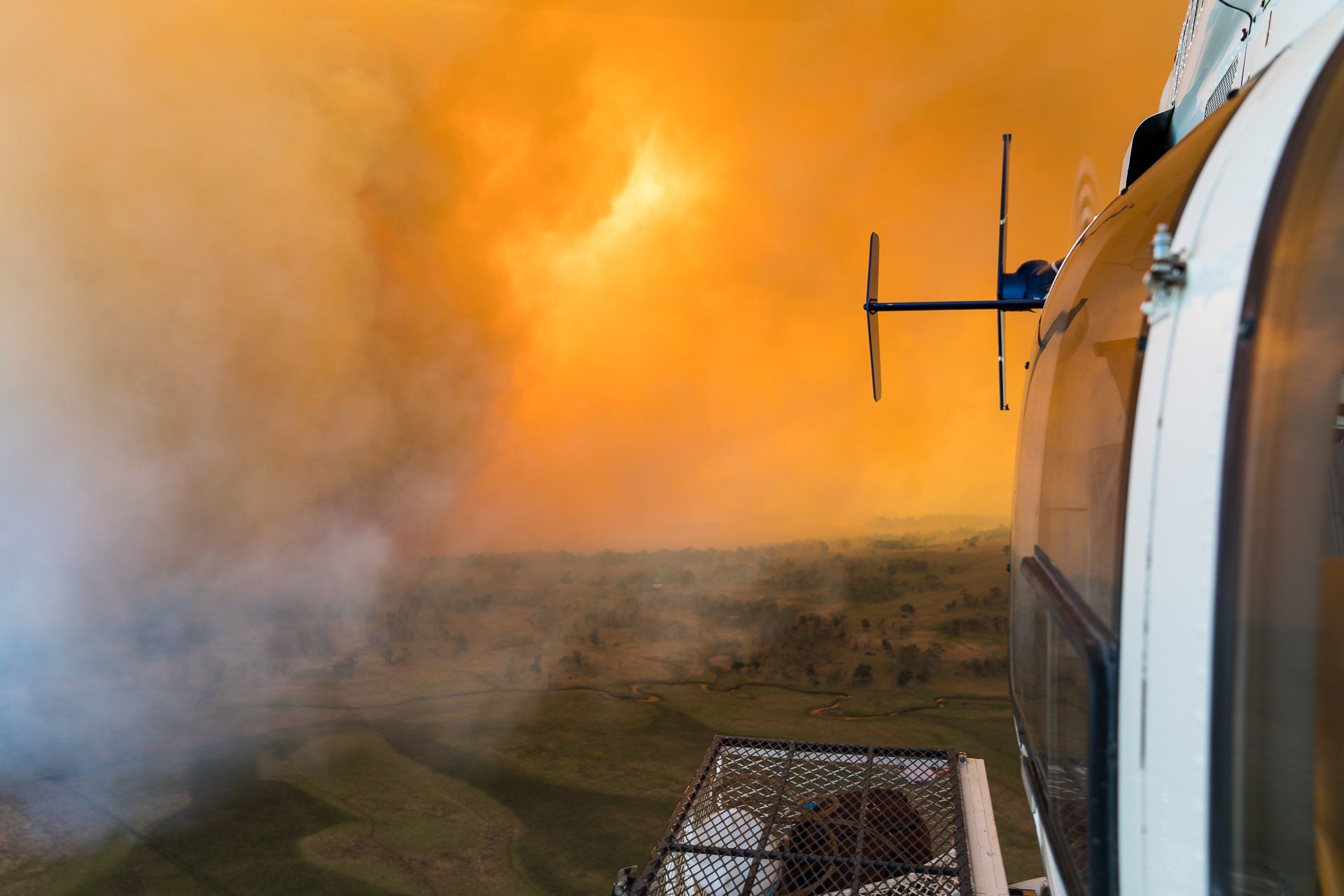 Helicopter flying near a wildfire with orange sky and smoky ground.