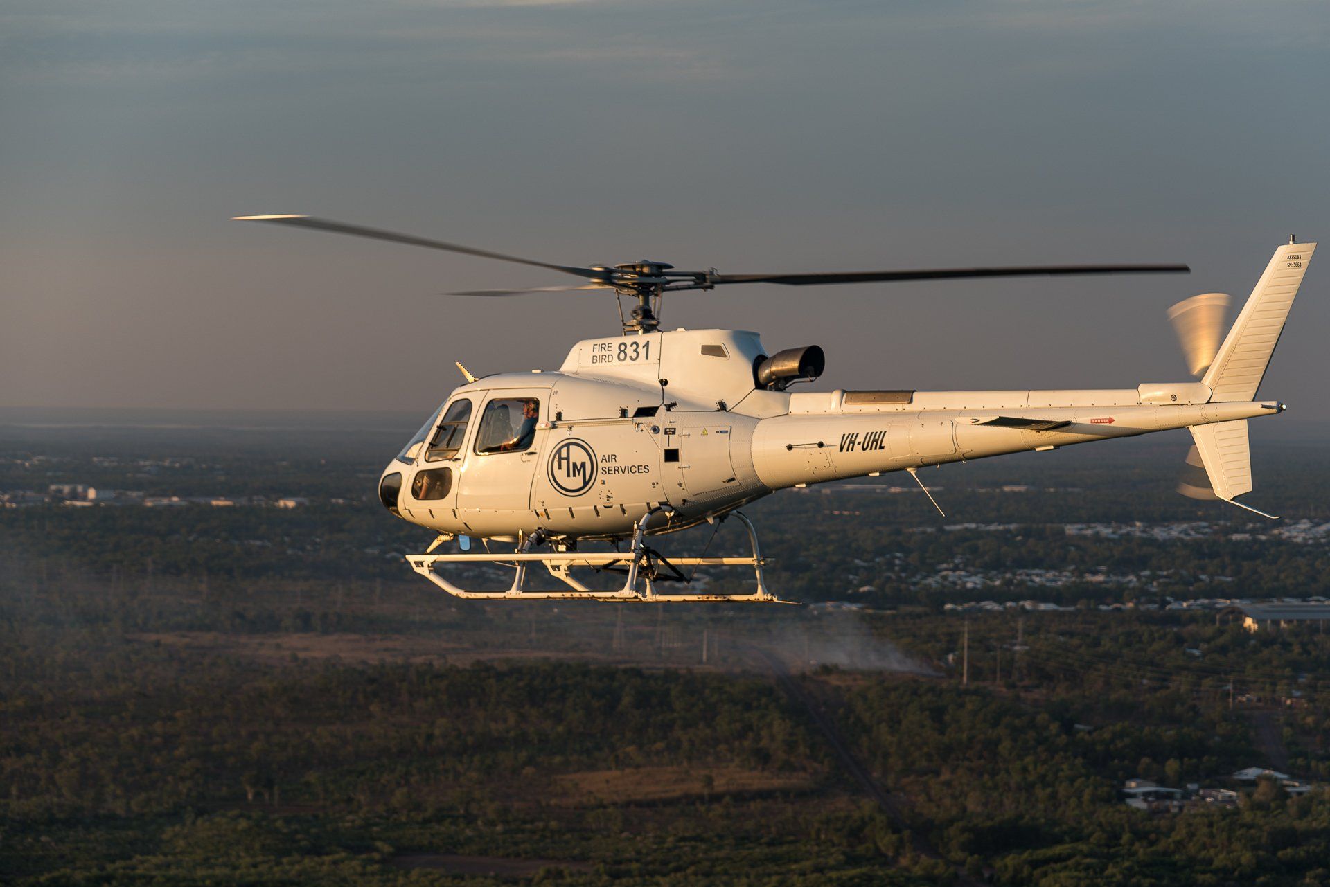 White helicopter flying over a forested area at sunset.