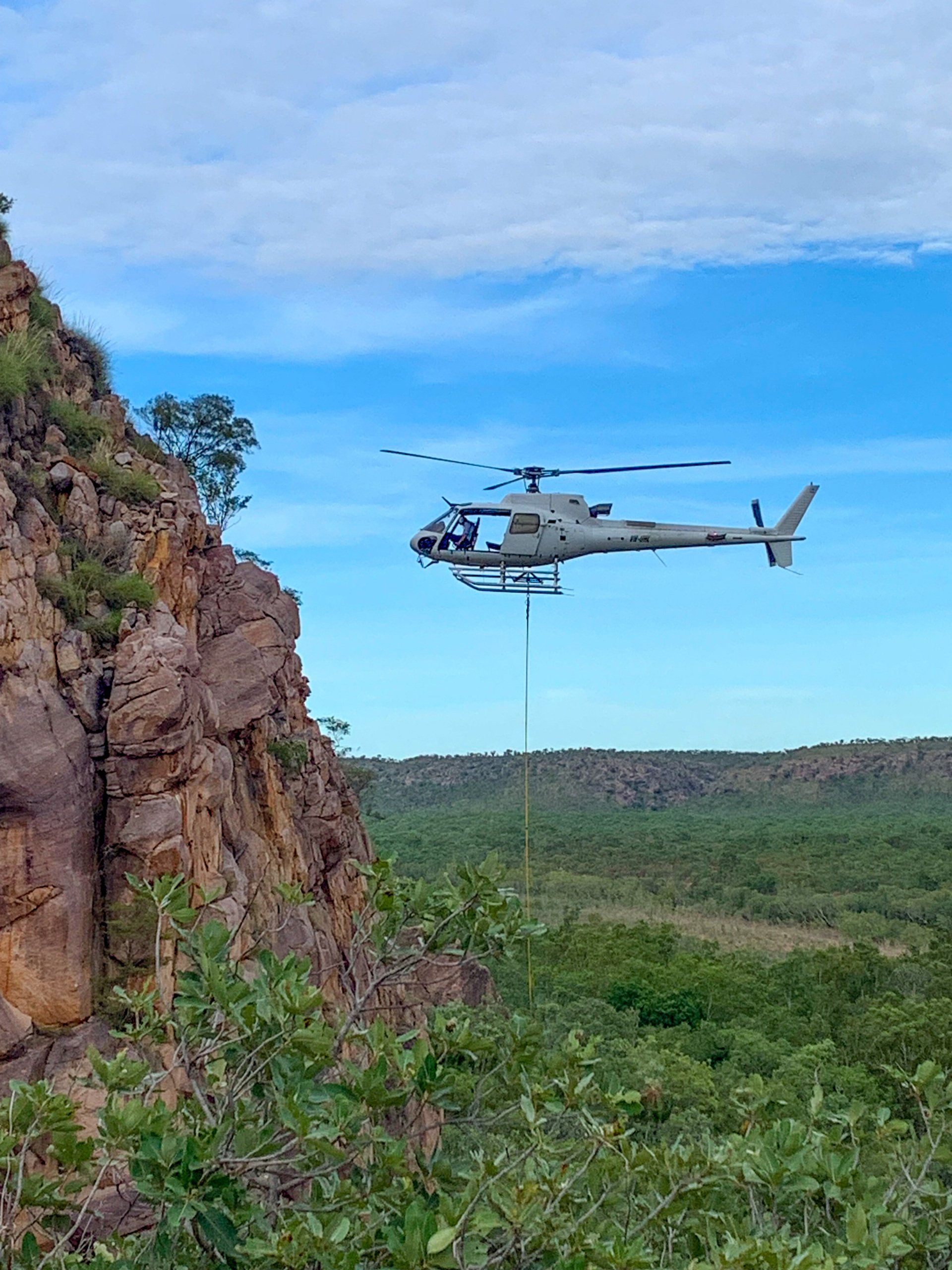 A military helicopter flies near a rocky cliff, over a forested landscape under a blue sky.