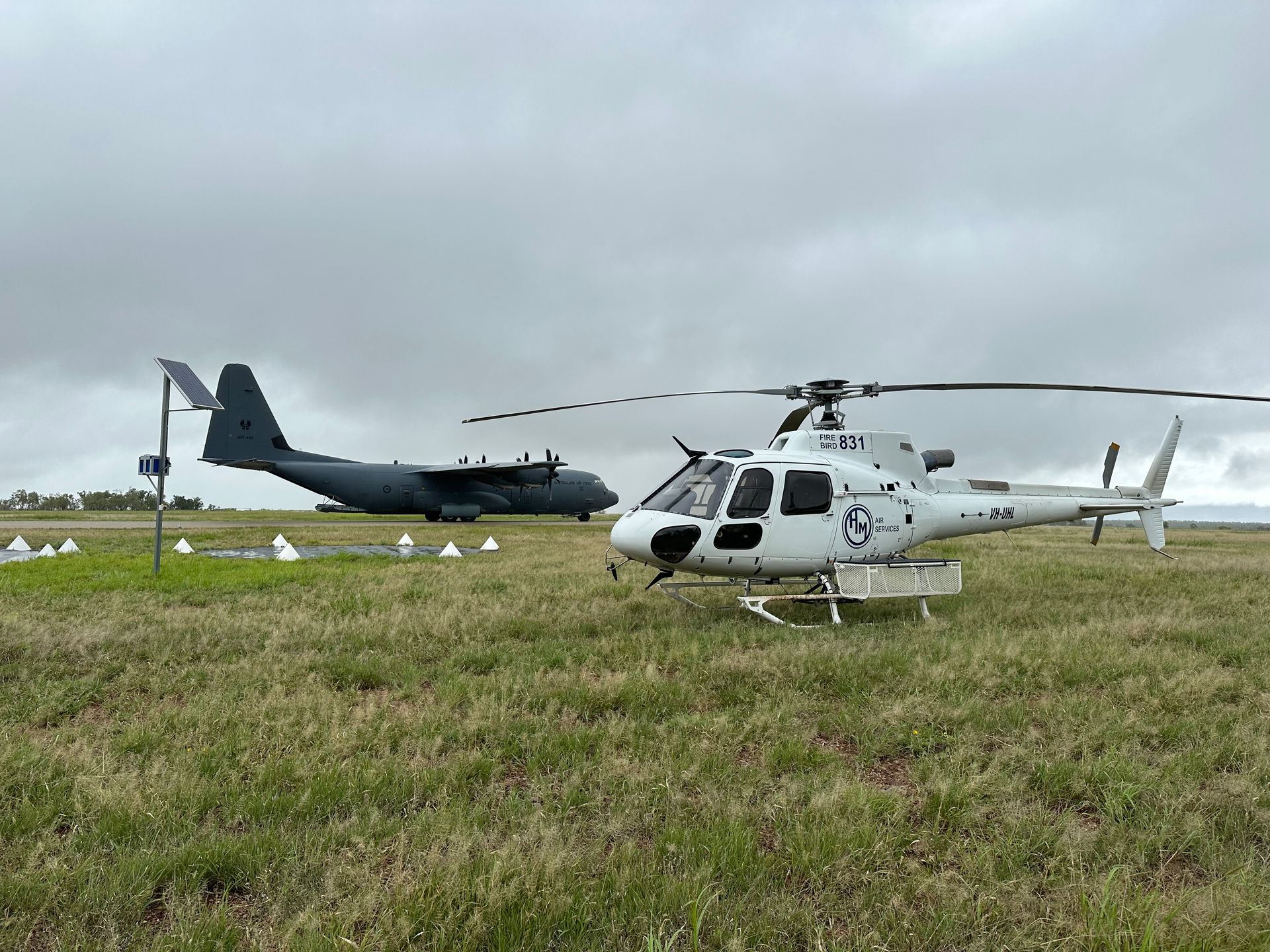 Helicopter parked on grassy airfield, C-130 in background under overcast sky.