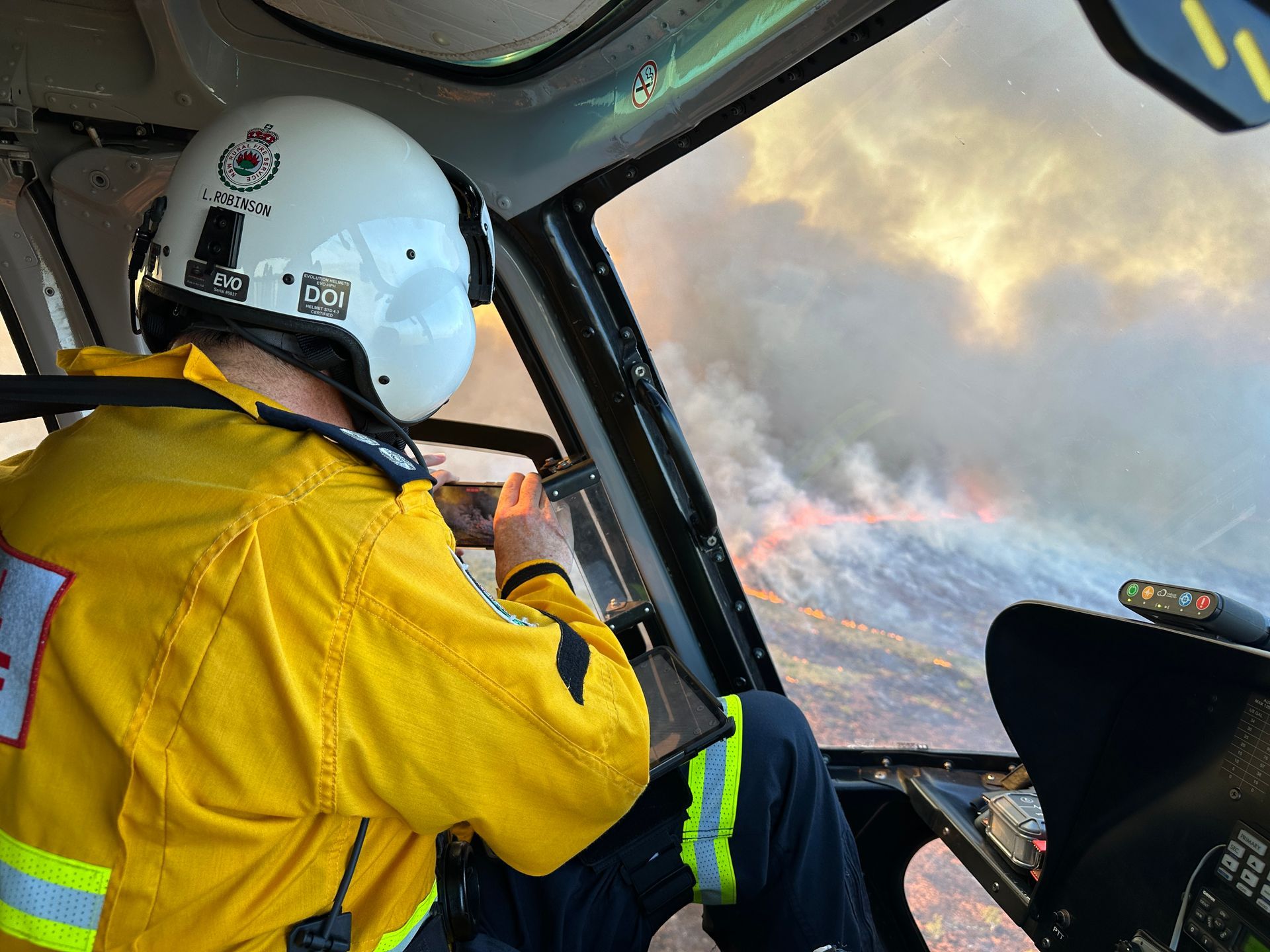 Helicopter cockpit view of firefighter aiming at a wildfire, wearing yellow jacket and helmet.