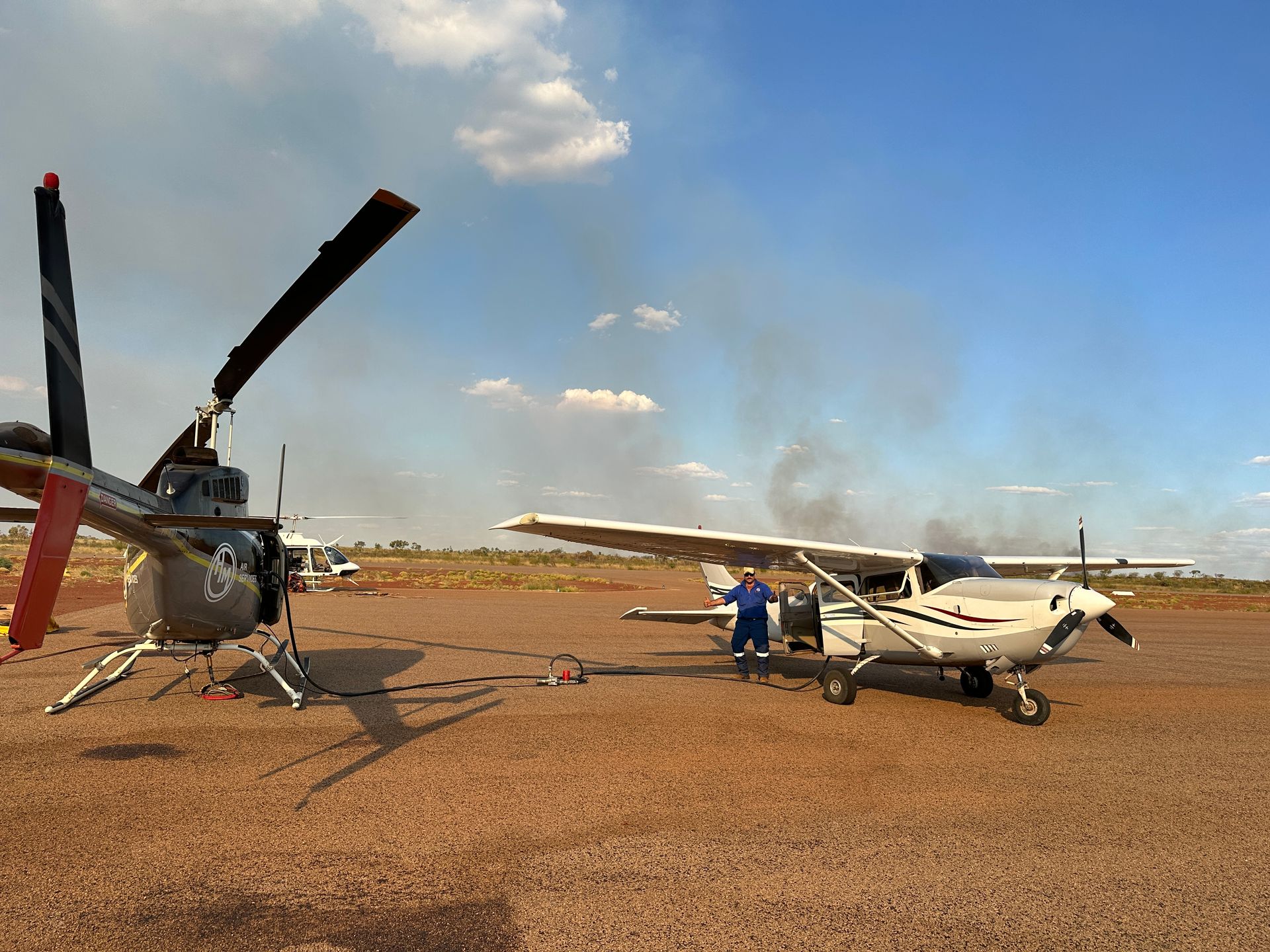 Helicopter and small planes on a dirt airstrip under a blue sky.  A person works on a plane.