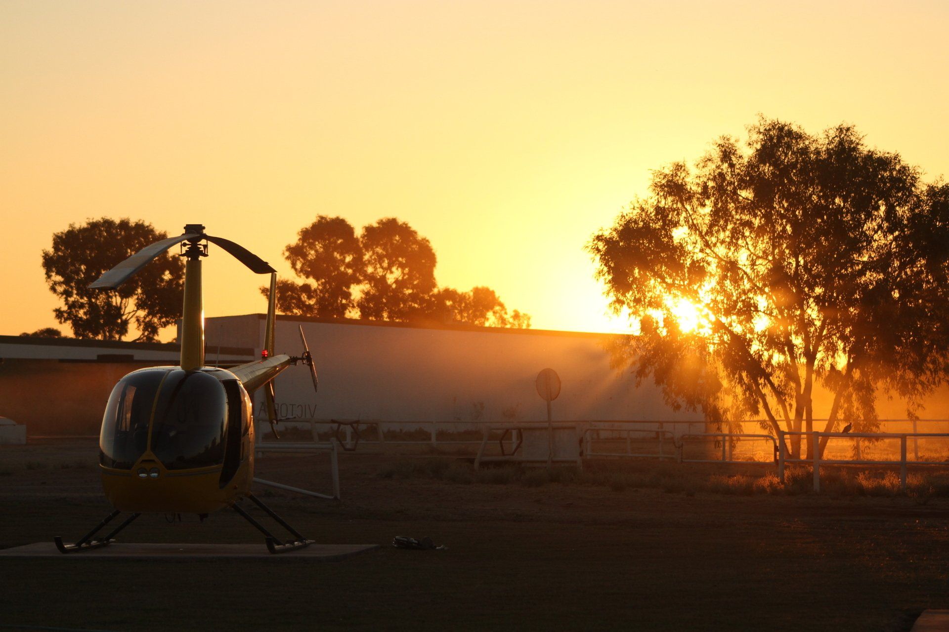 Yellow helicopter on the ground at sunset, with orange sky and tree silhouette.