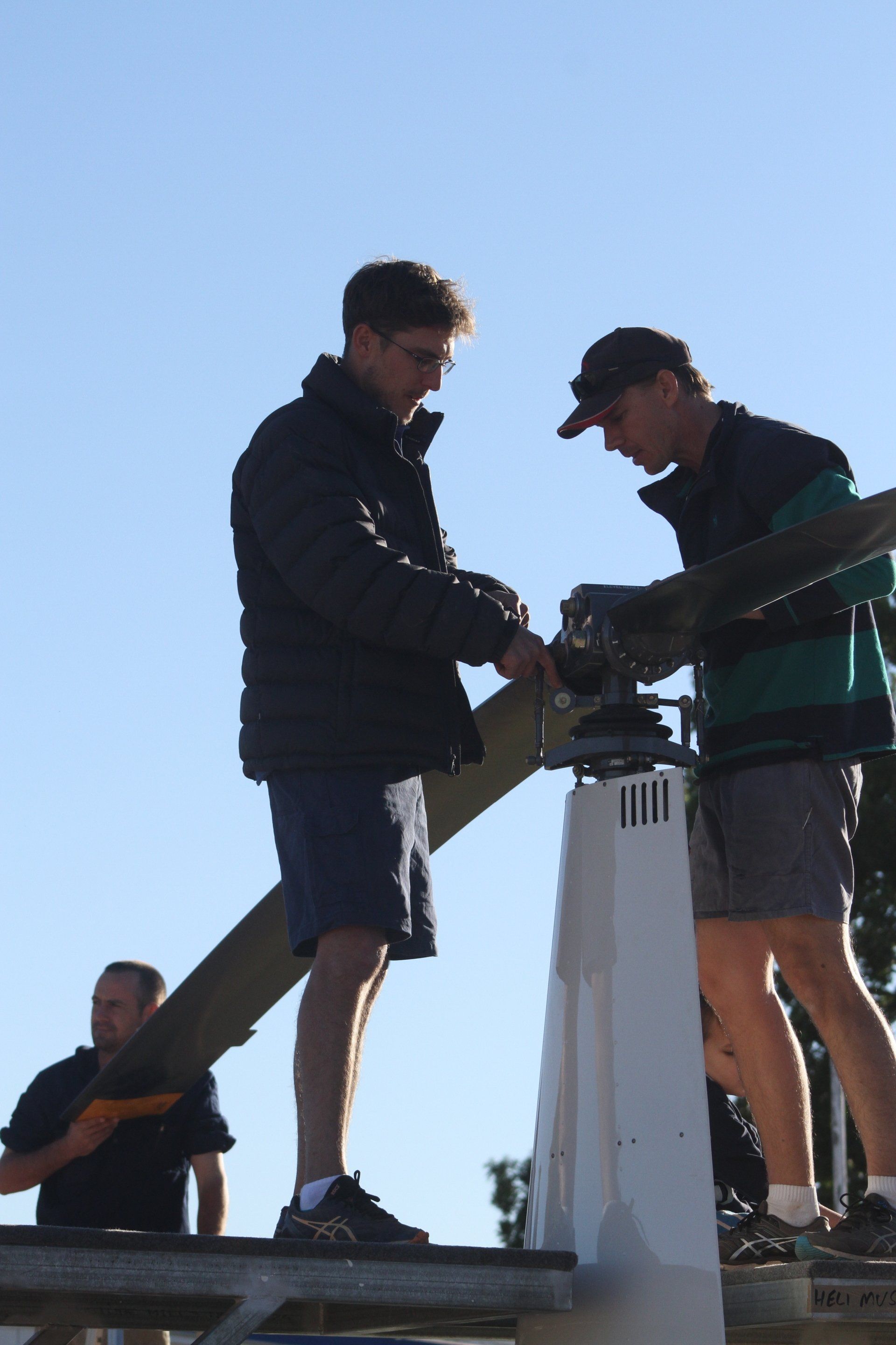 Two men adjusting a mechanism on top of a white structure, another man below holding a wooden beam, outdoors.