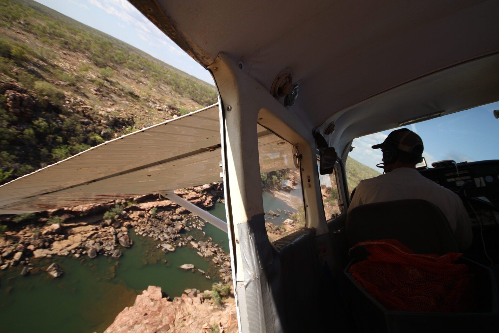 Pilot flying a small plane over a canyon. River below, rocks, wing visible, blue sky.