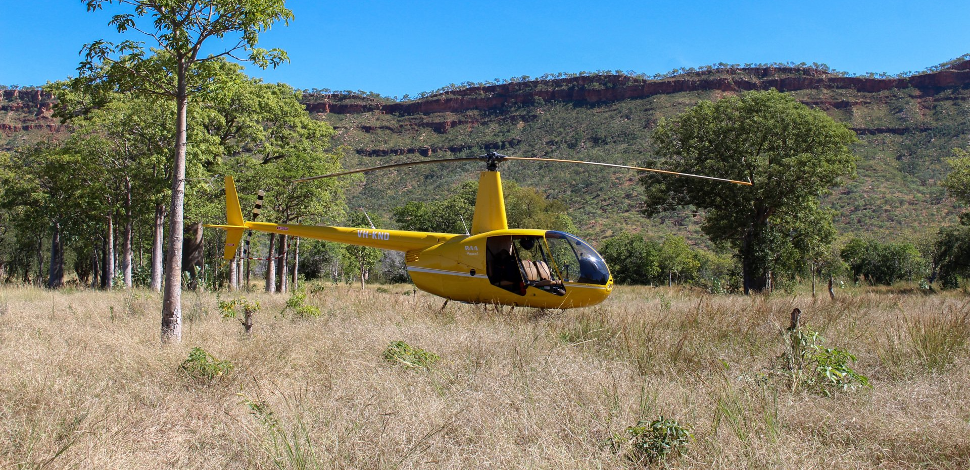 Yellow helicopter in field, trees in background, against mountains and blue sky.